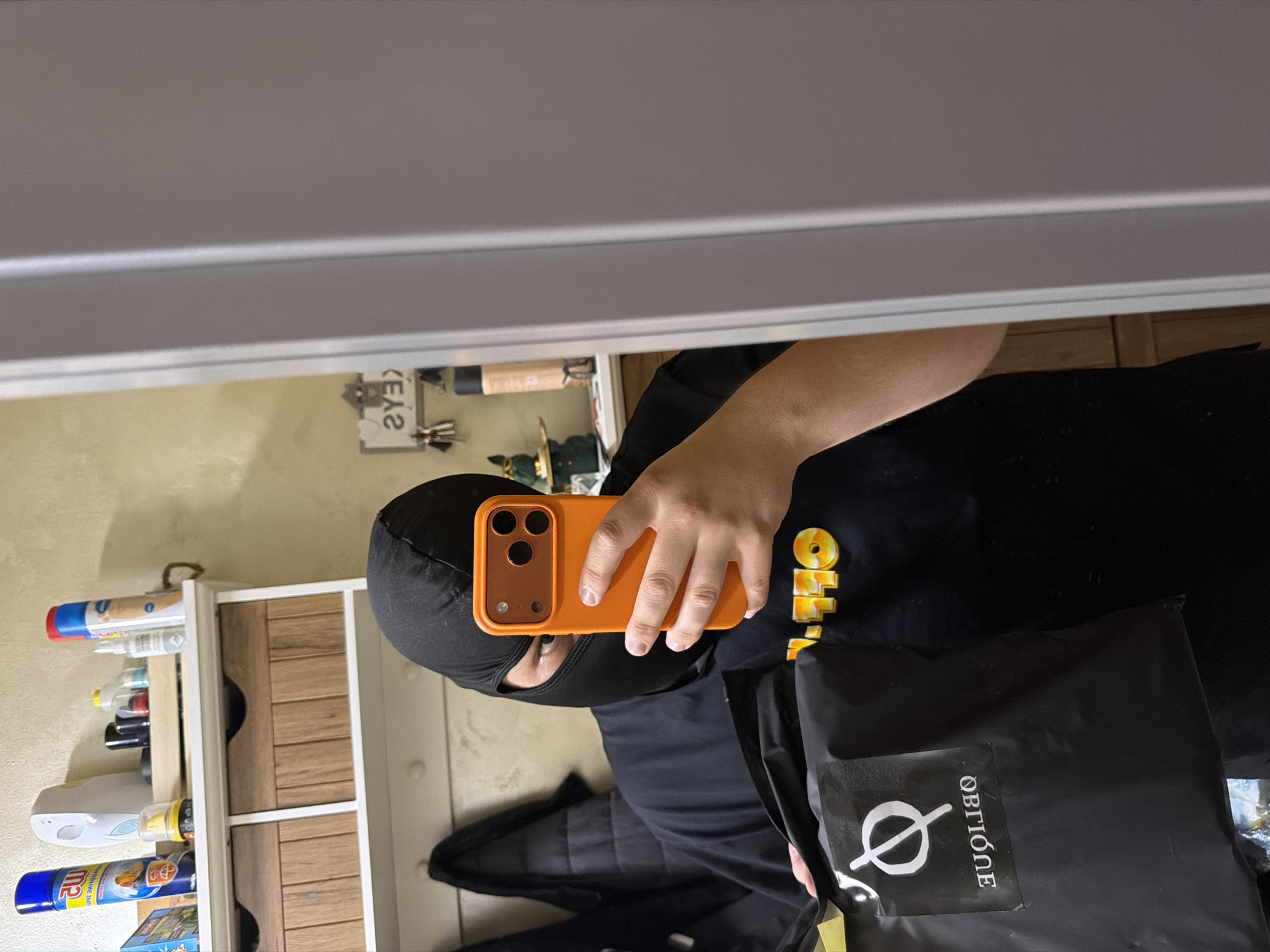 The image captures a mirror selfie taken indoors. The main subject is a person wearing a black balaclava, revealing only one eye. They are dressed in a black t-shirt with partially visible yellow text that reads "OFF-", likely part of a brand name. The person is holding an orange smartphone with a triple-lens camera setup in their right hand, using it to take the photo. A black bag with a distinctive white logo (a circle with a diagonal line through it, Ø) and the text "OBTIÓNE" is held in their left hand or draped nearby. A black jacket or similar garment is also visible.
The background shows a light-colored wall. To the person's left, a white shelving unit with wooden drawers or compartments holds various bottles and containers, possibly personal care or cleaning products. Above this unit, on the wall, hang what appear to be keys and fishing rods or similar tools. A small white label with mirrored and upside-down text, appearing as "2Y3)", is also visible. The top portion of the image reflects a grey overhead structure or ceiling. The setting suggests a casual, indoor environment, likely a room or hallway.