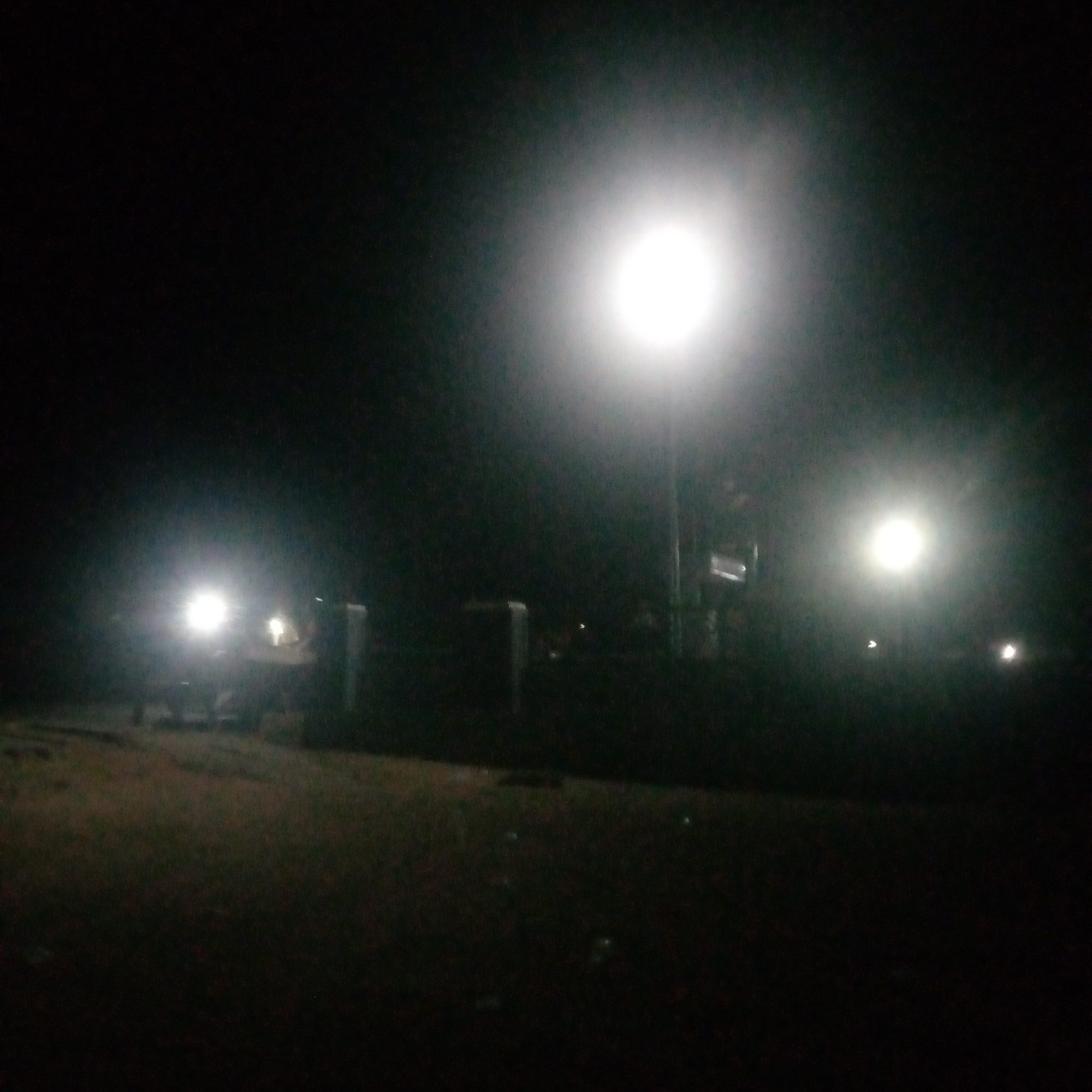 The image depicts an outdoor scene at night, likely in an urban or suburban area. A bright, artificial light source, possibly a streetlamp or floodlight, is prominently positioned in the upper center of the frame, casting a strong illumination that washes out details in its immediate vicinity. To the left of this central light, a fainter, second light source is visible, and further to the left, another diffused light is present, suggesting the presence of vehicles or other lighting.
The ground in the foreground appears to be unpaved, possibly a dirt or gravel path, with some patchy vegetation visible. In the mid-ground, there are some dark, indistinct structures that could be fences, walls, or low buildings, partially silhouetted against the illuminated background. The overall environment is dark and lacks clear features due to the nighttime setting and the intensity of the artificial lighting. No people are clearly discernible in the image. The location is stated to be in the city of Unknown, Unknown.