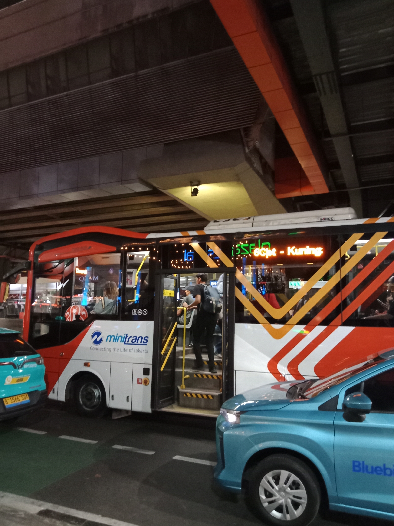 The image shows a street scene in South Jakarta, Indonesia, likely at night. In the foreground, a blue car with "Bluebird" branding is visible. Behind it, a red and white Minatrans bus is stopped. The bus displays a digital sign with orange text that reads "Fiselfict - Kuning". Several people are visible on the steps of the bus, either boarding or disembarking. The environment appears to be urban, with elevated structures and lighting visible in the background, suggesting an area with overpasses or a transportation hub. The overall lighting suggests it is evening or night.