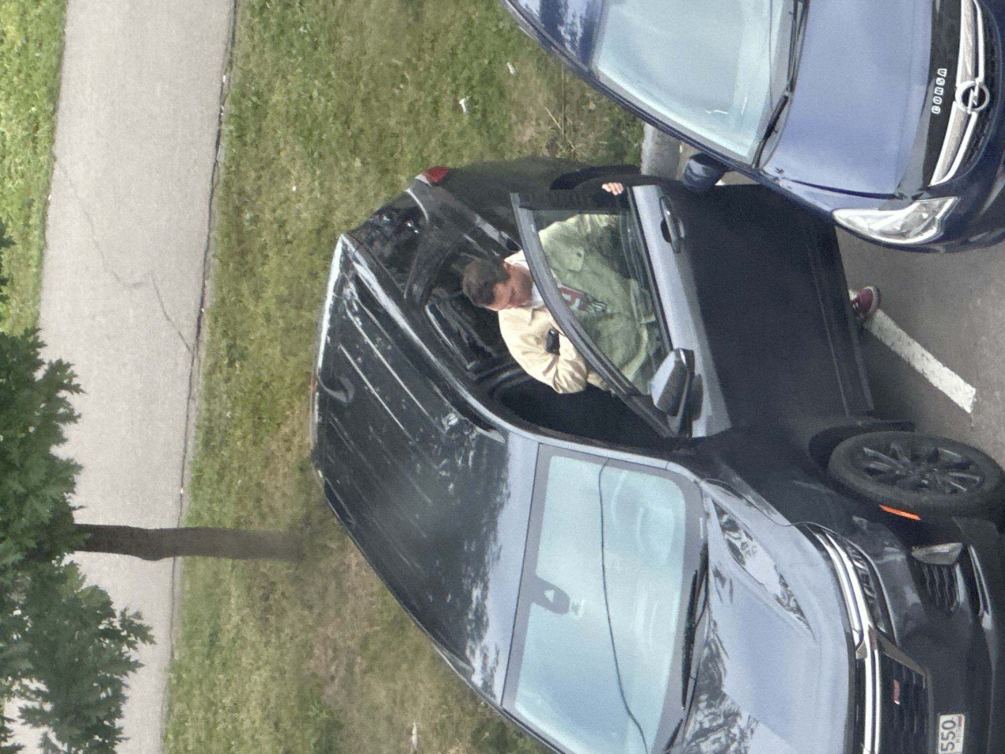 An overhead, eye-level, medium shot captures a man emerging from the driver's side of a black SUV, parked on a paved surface adjacent to a grassy area with a tree. The man is wearing a light-colored jacket over a t-shirt, and red sneakers. He is reaching into the vehicle. A blue car is parked to the right of the black SUV, with its hood visible. The license plate on the front of the black SUV is partially visible, showing the numbers "550". The image appears to be taken in the city of Podolsk, Russia.