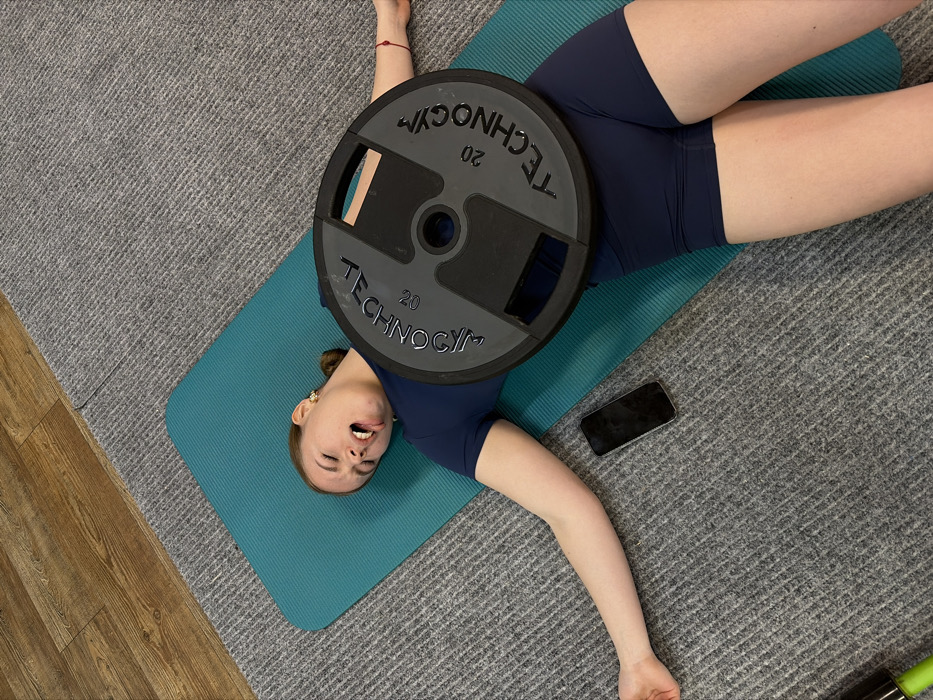 A female individual lies supine on a teal exercise mat in an indoor gym or workout space. She is dressed in a dark blue short-sleeved athletic top and matching shorts. Her eyes are closed, and her mouth is open. A dark grey Technogym weight plate, marked "20" on its surface, rests across her lower chest and abdomen. Her arms are extended to her sides, with hands open and palms facing upwards. A black smartphone is placed screen-down on the grey carpet near her left hand. A green object is partially visible in the bottom-left corner of the image. The flooring consists of grey carpet, transitioning to wooden floorboards in the upper left. The precise location within the city of Chaikovsky, Russia, is not identifiable from the image.