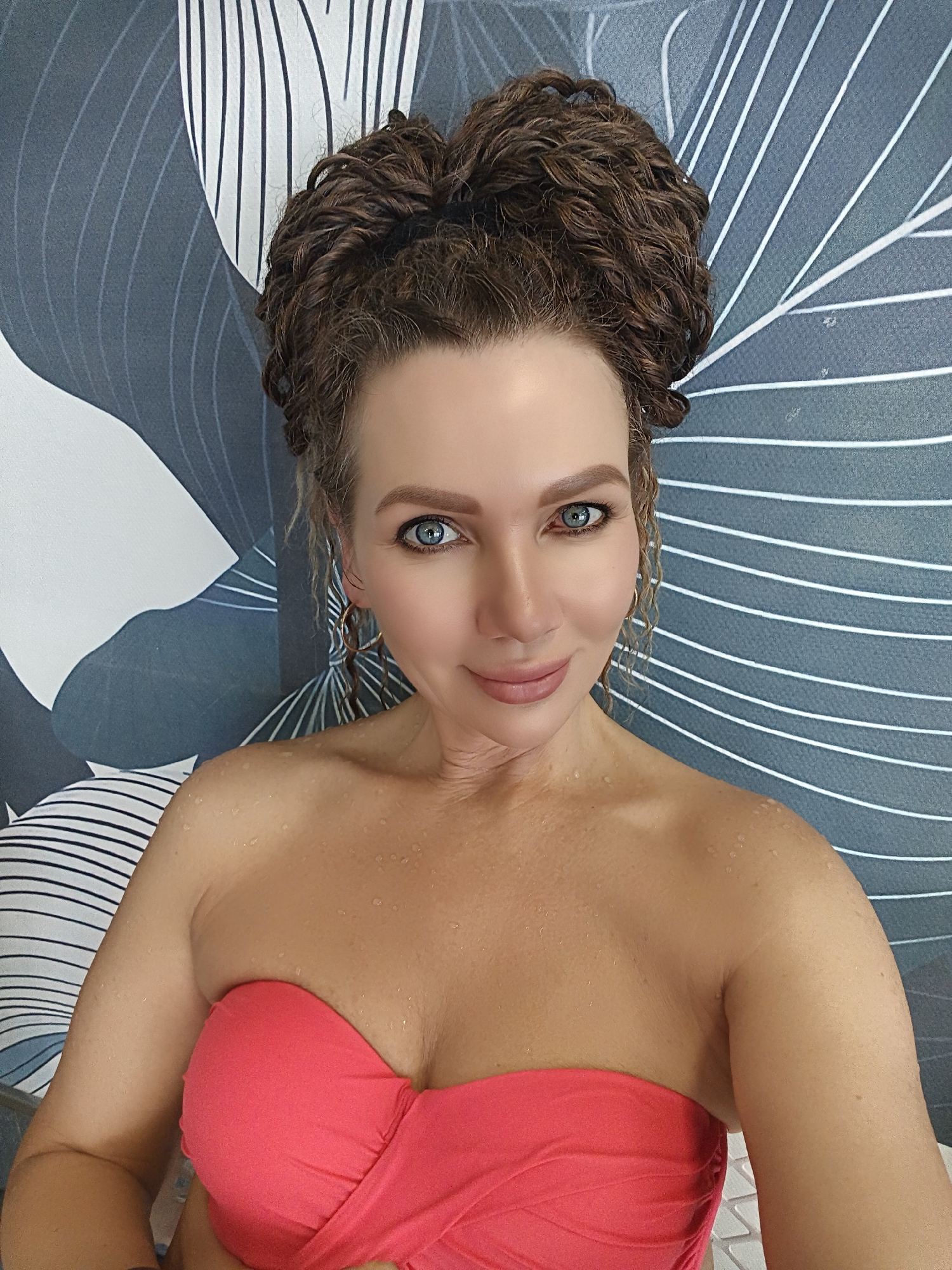 A woman with curly brown hair styled in a high bun and bright blue eyes is shown from the chest up, looking at the camera. She is wearing a strapless coral-pink bikini top. Her skin is wet, with small water droplets visible on her shoulders and décolletage. She has on hoop earrings and her eyebrows are well-defined. The background is a shower curtain with a pattern of grey and white leaf-like shapes and thin white horizontal stripes. The lighting appears to be from an overhead source. The image was taken in Chaikovsky, Russia.