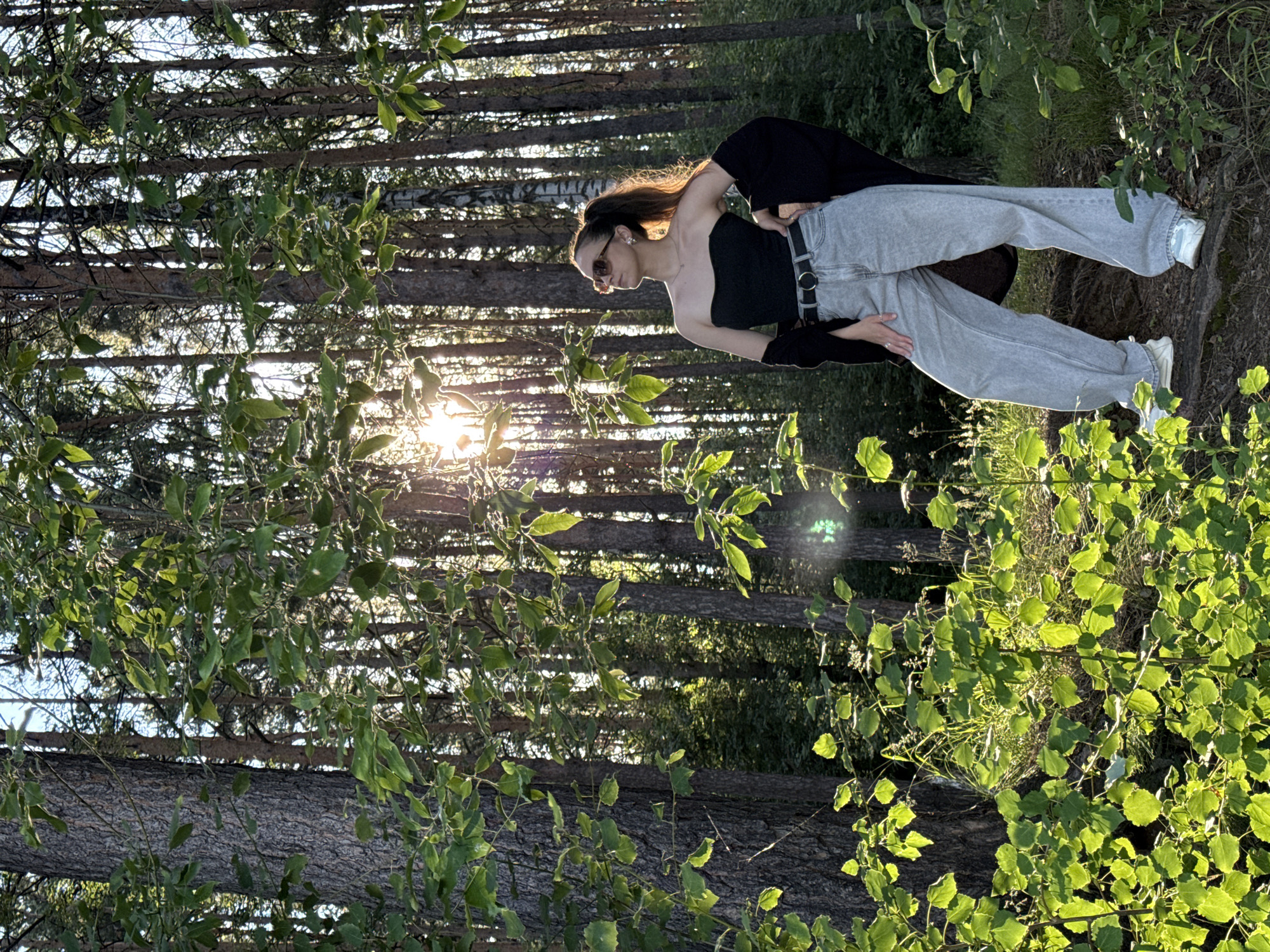A young woman stands in a sun-dappled forest, her body turned slightly to the right as she looks upwards and away from the camera. She has a ponytail and wears sunglasses, a black strapless top, a black open cardigan draped over her shoulders, light-wash wide-leg jeans, a black belt, and white sneakers. The sun's rays filter through the tall trees, creating a dramatic lighting effect on the leaves and the woman. The location is identified as a forest in Chaikovsky, Russia, with dense trees and undergrowth. The ground is covered with dirt and fallen leaves, with some rocks and roots visible. The overall atmosphere is natural and serene, with the light playing a significant role in the composition.