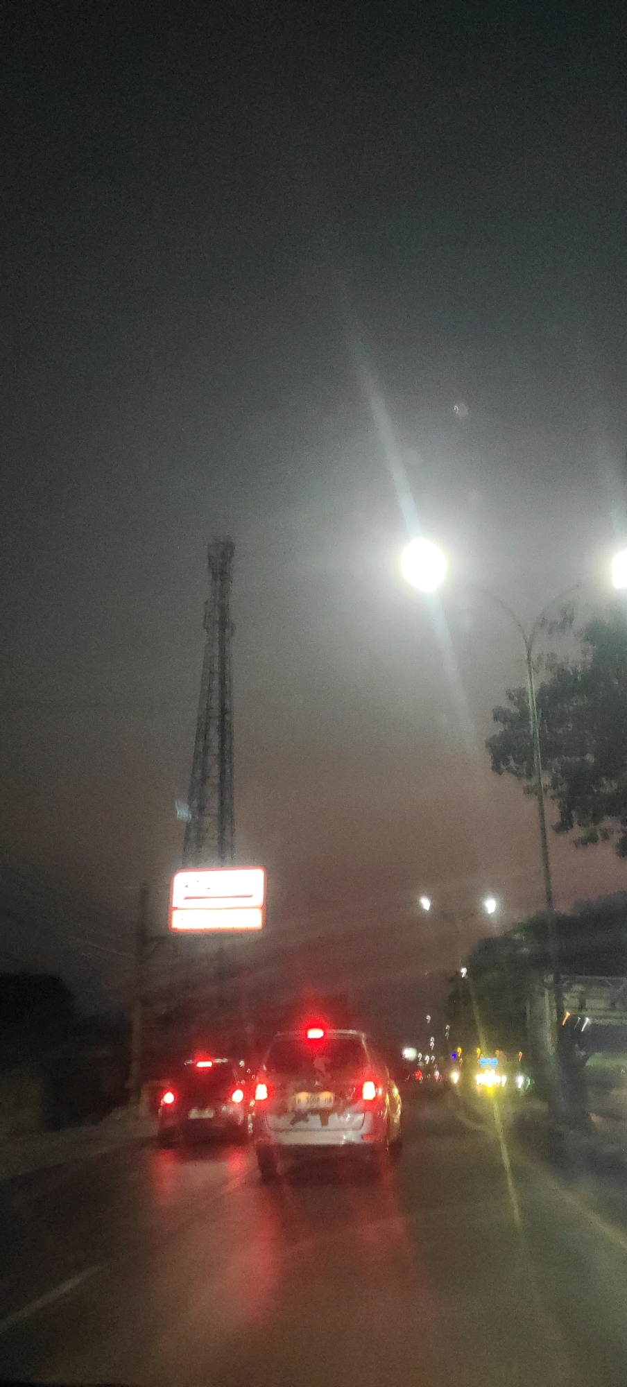 The image depicts a nighttime street scene in Bolo Krajan, Indonesia. The sky is overcast and dark, with a bright street light illuminating the scene on the right side. A tall communication tower stands prominently in the distance on the left, with a lit billboard sign below it. Several cars are visible on the wet road, their taillights glowing red. The foremost car is a white SUV with a license plate that appears to read "B 1008 HA." The overall impression is a slightly foggy, atmospheric night drive.