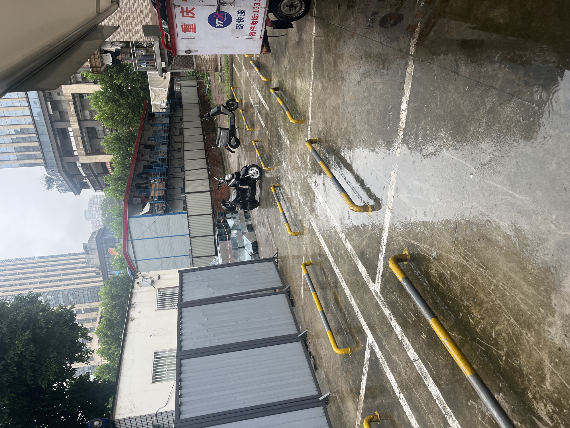 The image depicts an urban outdoor area, likely a parking or loading zone, on a wet and overcast day in Yuzhong District, Chongqing, China. The concrete ground is extensively wet, showing numerous concentric ripples from recent or ongoing rainfall. White, faded lines delineate parking sections on the pavement. Several U-shaped metal barriers, painted yellow and grey, are anchored into the ground, arranged in rows. Two dark motor scooters, one with "F90" visible on its side, are parked between these barriers in the midground. On the far right, the rear section of a white delivery truck or three-wheeled vehicle is partially visible, displaying the characters "重 庆" (Chóngqìng), a "YT express" logo, and Chinese text for "send express" along with a phone number.
To the left, a series of tall, grey corrugated metal temporary fences with dark frames form a barrier. Behind this, a white single-story building with two barred windows is visible, partially obscured by green tree foliage. In the mid-background, a red-roofed, blue-sided structure with items resembling clothes hanging from it stands. Further back and to the right, a multi-story brick building with balconies displays laundry hanging to dry. The background is dominated by a tall, modern high-rise building with extensive fenestration, and other urban structures are visible through the hazy, grey sky. No individuals are present in the scene.