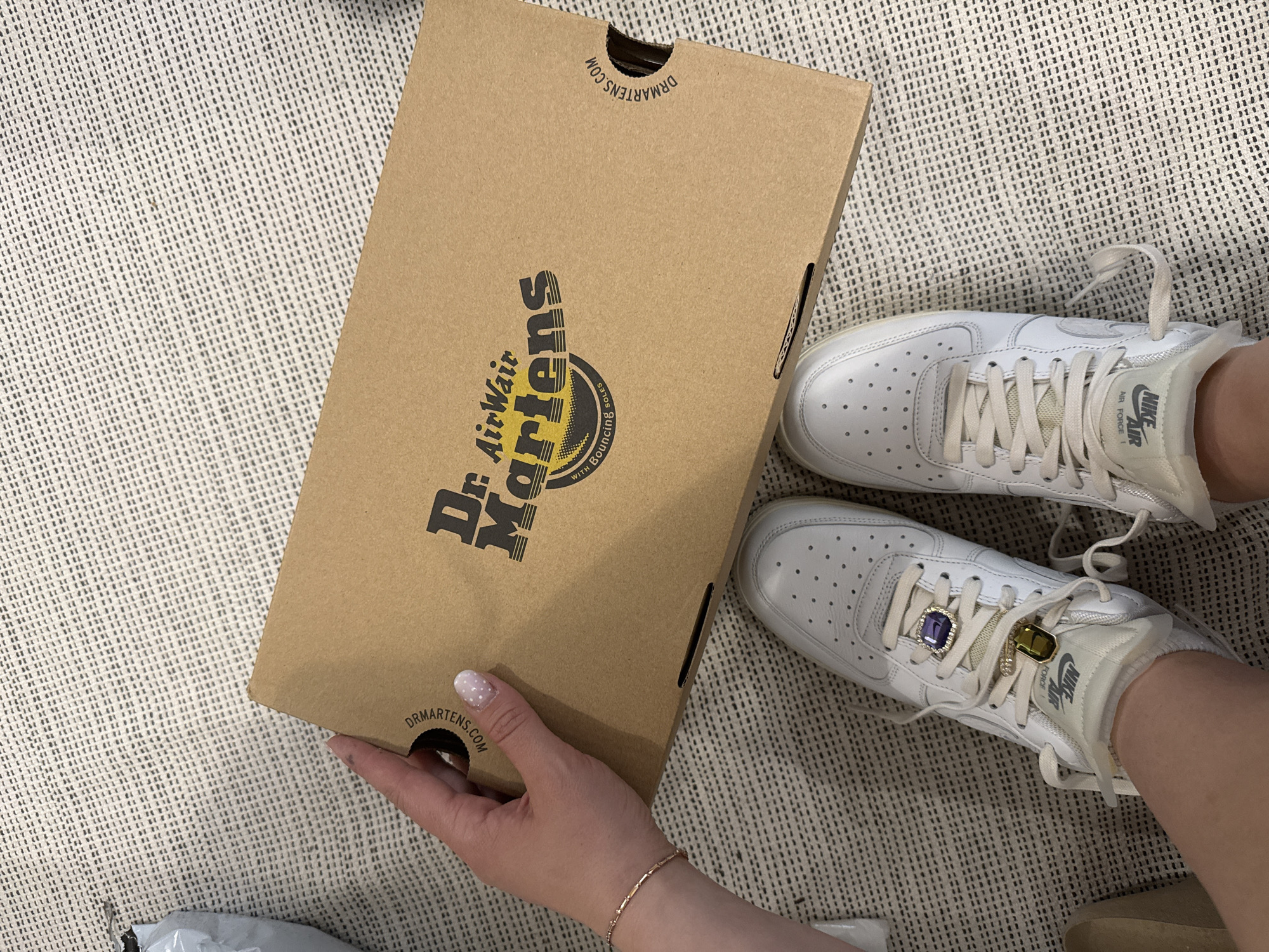 The image captures an overhead view of a person indoors, likely in Hainburg an der Donau, Austria, holding a Dr. Martens shoebox. The person is wearing a pair of white Nike Air Force 1 sneakers, which feature decorative jewel-like lace charms, one purple and one green/yellow. A hand with light-colored, polka-dotted nail polish and a delicate bracelet is seen holding the cardboard shoebox.
The shoebox itself is a light brown kraft paper color, displaying the "Dr. AirWair Martens" logo prominently in black, along with the phrase "WITH BOUNCING SOLES" and the website "DRMARTENS.COM" printed upside down on the top flaps. The person's feet, clad in the sneakers, rest on a light-colored, textured rug or carpet. The tongue of the sneakers clearly shows "NIKE AIR FORCE 1" branding. The scene suggests a casual moment, possibly an unboxing or showcasing of new items.