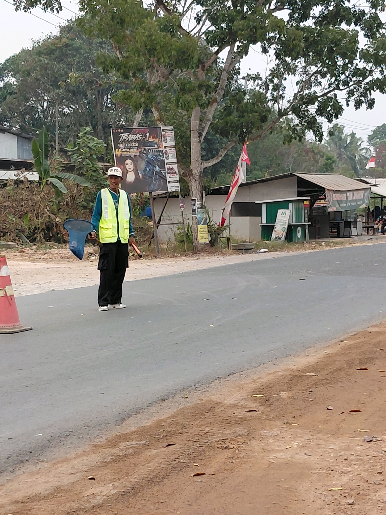 A man wearing a yellow safety vest and a white cap is standing on a paved road, holding a blue net and an orange baton. He is positioned in front of a large poster advertising "TRABAS 1", which features images of vehicles and people. To his right, there are several small buildings with awnings and some vegetation. In the background, trees and more buildings are visible. The road surface appears worn, and there is a traffic cone to the left of the man. The scene suggests an outdoor setting, possibly in a rural or semi-urban area in Persiapan Mulya Raya, Indonesia, given the context.