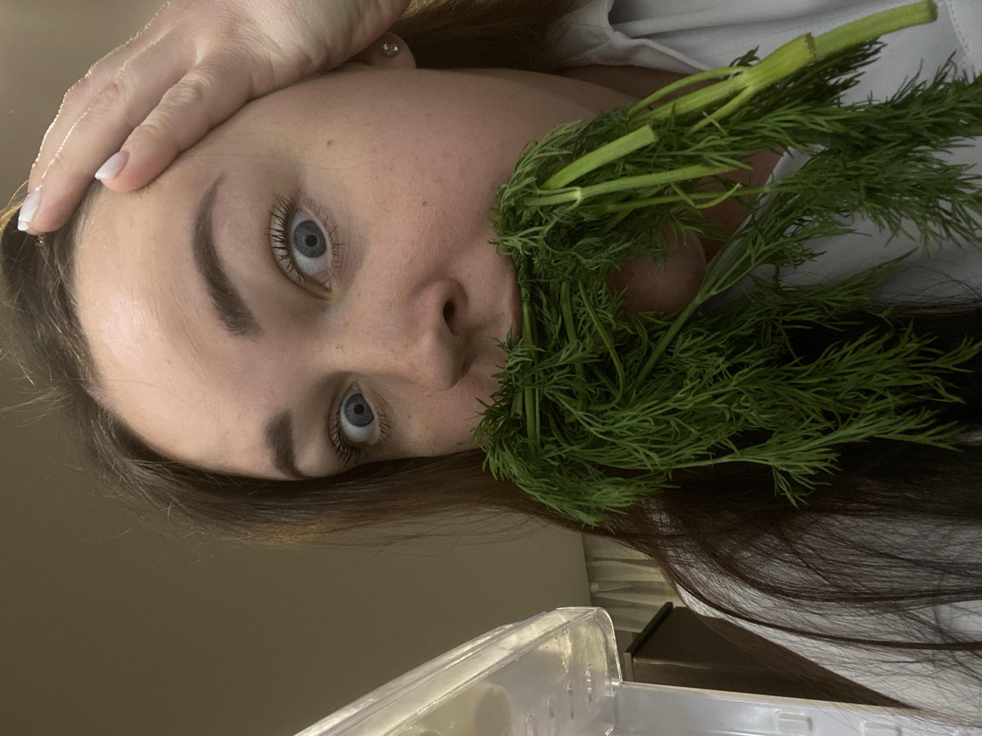 A woman is holding a bunch of dill to her face. She is looking at the camera with her left eye. Her left hand is resting on her forehead. She has brown hair and blue eyes. A clear plastic container is visible in the lower part of the frame.
The location of the photo is not evident. There is no basis to assume it was taken in Sochi, Russia.