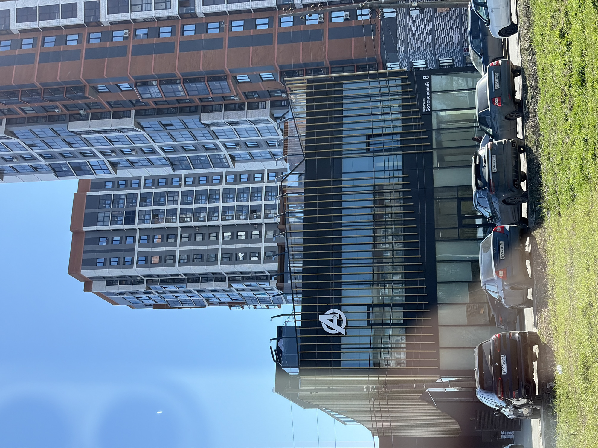 Here is a detailed description of the image:
**Scene:**
The image appears to be a daytime photograph taken outdoors, showcasing a street view with modern buildings and parked cars. The angle seems to be slightly upward, capturing the height of the structures.
**Objects:**
* Several multi-story buildings dominate the background. They are characterized by a modern architectural style, with glass windows and contrasting façade elements in shades of gray, brown, and black.
* A smaller building with a black facade and gold decorative beams features a large logo that resembles an inverted “Q” or a stylized “A”. The street address is “Переулок Ботеневский 8” (Botenevskiy lane 8)
* Parked along the street are a line of cars, including a black BMW X7, a gray Ford Focus, and a white sedan. The license plates of the cars show the region code "18", indicating the Udmurt Republic, Russia.
* A patch of green grass lines the edge of the street, bordering the parked cars.
**Environment:**
* The sky is clear blue with some brightness.
* The overall light suggests a sunny day, casting shadows from the buildings and cars.
**Location:**
The street address "Переулок Ботеневский 8" specifies that this location is Botenevskiy Lane 8. Given the region code from the license plates, the image captures the modern architecture in the city of Izhevsk, Russia.