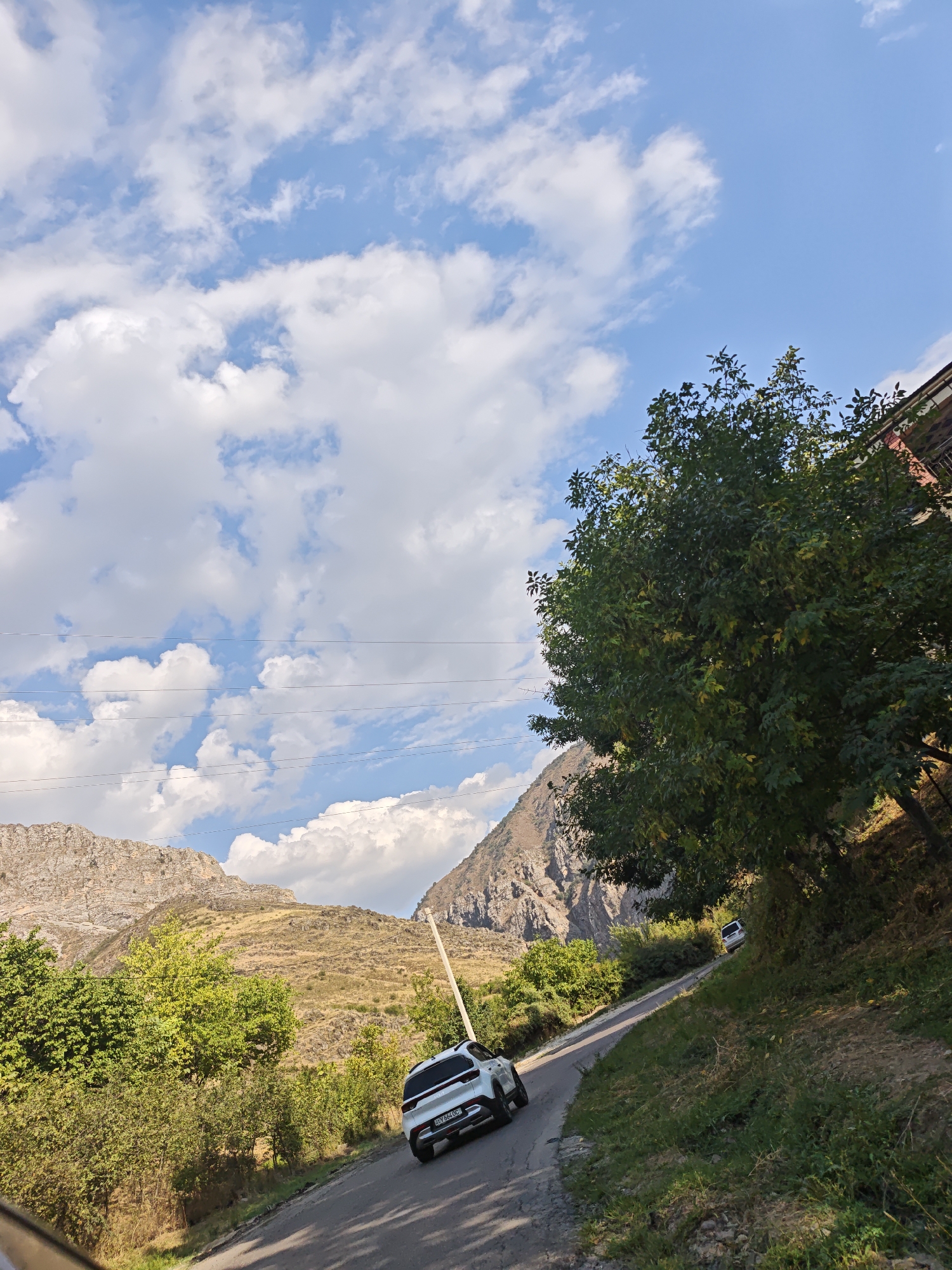 The image depicts a scenic view on a sunny day in Brichmulla, Uzbekistan. A paved road winds through a mountainous landscape, with lush green trees and vegetation lining its sides. In the foreground, a white SUV is seen driving away from the camera. Further up the road, another white vehicle is visible in the distance. The sky is a vibrant blue, dotted with fluffy white clouds. The overall impression is one of a peaceful and picturesque natural setting, characteristic of the region.
