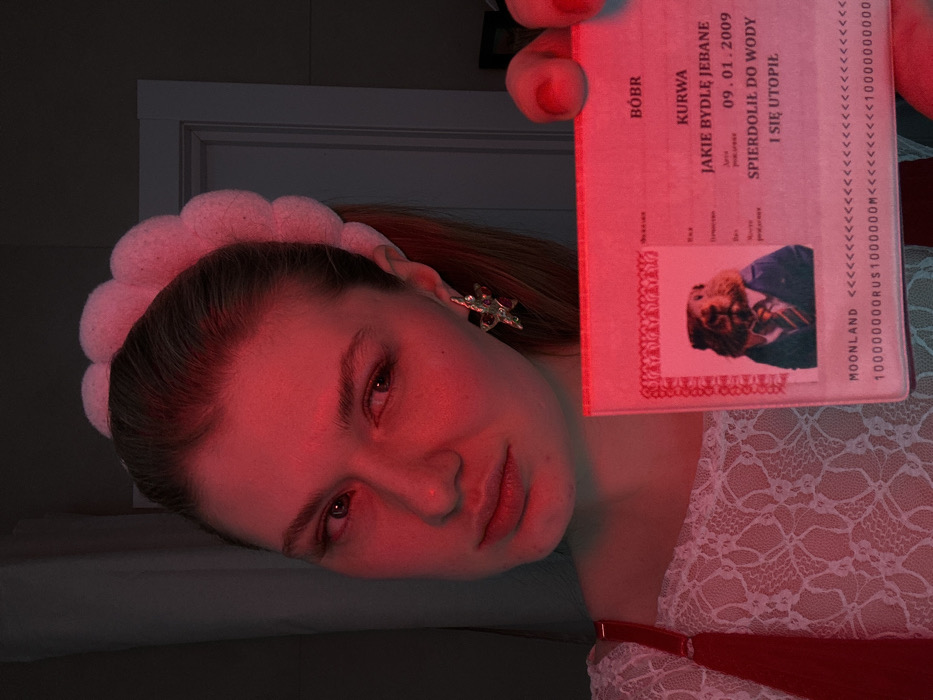 A person illuminated by a red light holds up a Polish ID-like document. The document contains phrases in Polish, including "BÓBR," "KURWA," and "JAKIE BYDLĘ JEBANE." It has an official format with a photo of a man in a suit, a date, and text that translates to "drowned in water." The bottom includes the word "MOONLAND" and a sequence of "<<" and numbers followed by "RUS" and "M". The person holding the document is wearing a headband and a white lace top.