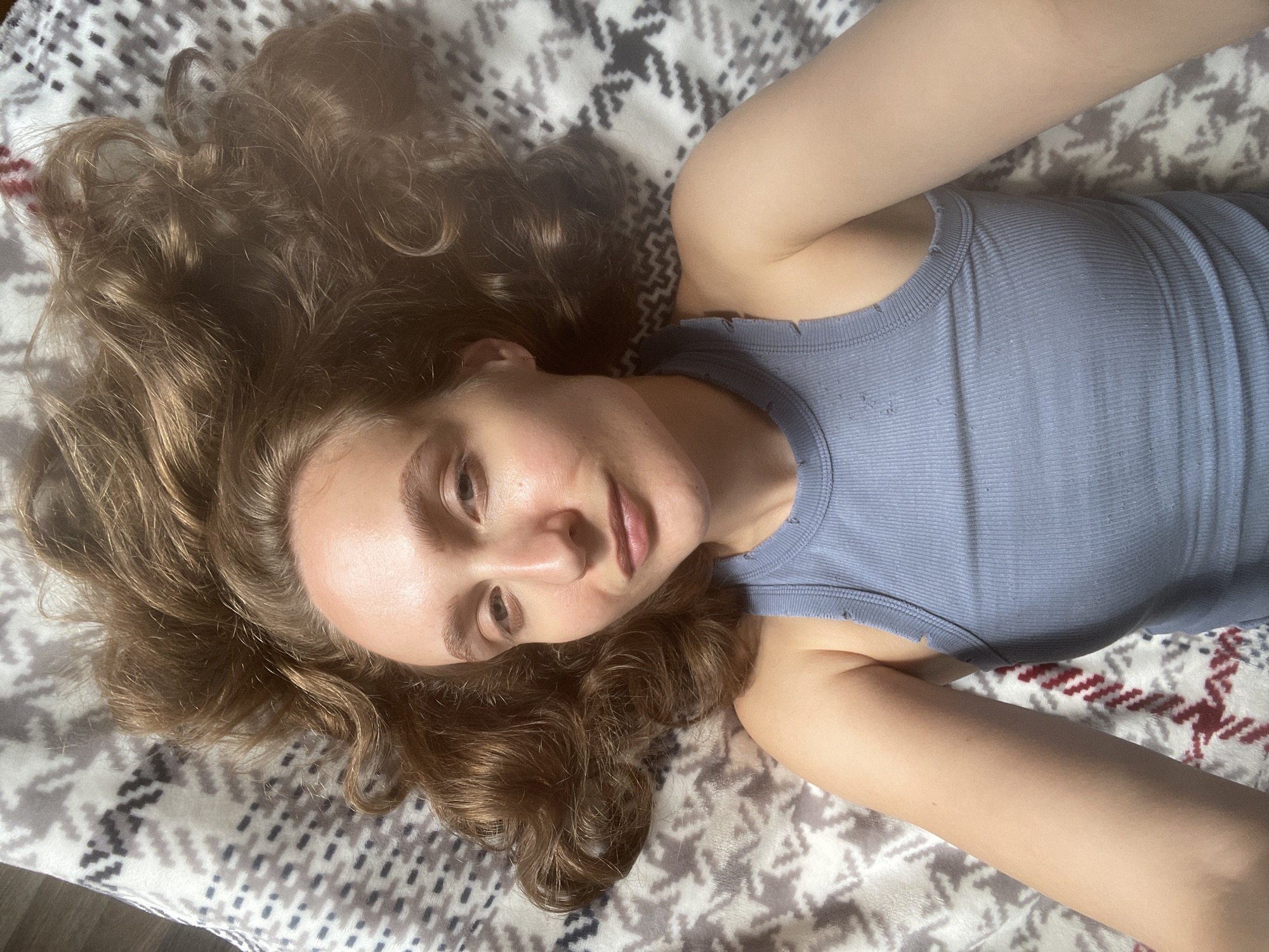 A young woman with light brown, wavy hair is lying on her back on a patterned blanket. She is wearing a light blue ribbed tank top. Her head is tilted slightly to her right, and her eyes are open, looking directly at the camera. The blanket has a gray and white geometric pattern. The scene appears to be indoors, possibly in a bedroom, given the presence of a blanket. There is no indication of the location being the city of Sharkan, Russia.