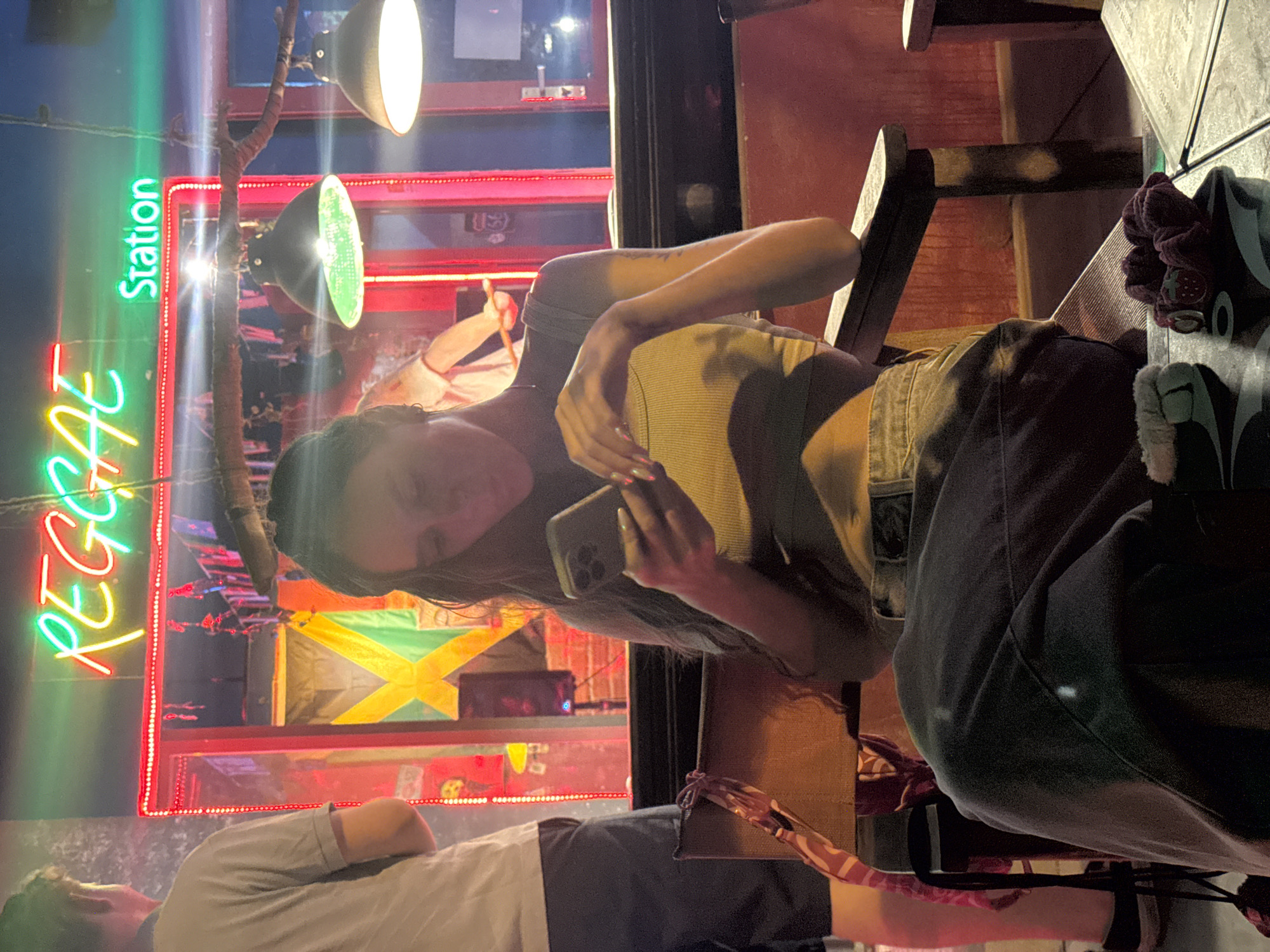 A woman is sitting at an outdoor bar in Đà Nẵng, Vietnam, holding a cell phone. The "Reggae Station" sign is visible behind her. A man sits across from her. The restaurant has bright red and green neon lights and the Jamaican flag.