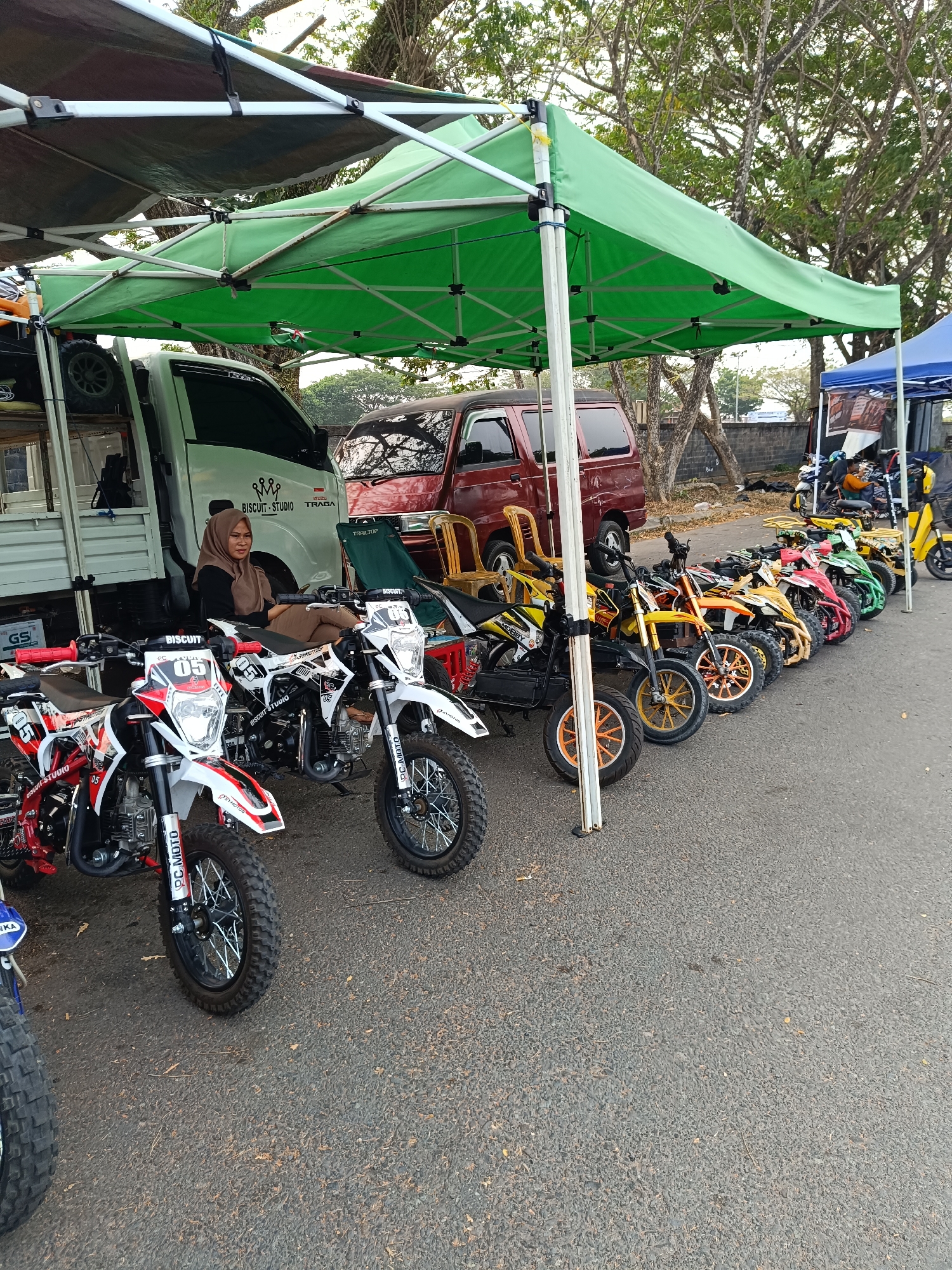 The image depicts an outdoor scene in Kayu Agung, Indonesia, likely a market or exhibition area. Several small dirt bikes and ATVs are lined up under a green canopy tent. A woman wearing a hijab is seated on one of the motorcycles, looking towards the camera. A white truck is parked behind the motorcycles, and a red van is visible to the right of the truck. The ground is paved with asphalt, and trees with dense foliage line the background. The overall impression is one of an organized display or sale of recreational vehicles.