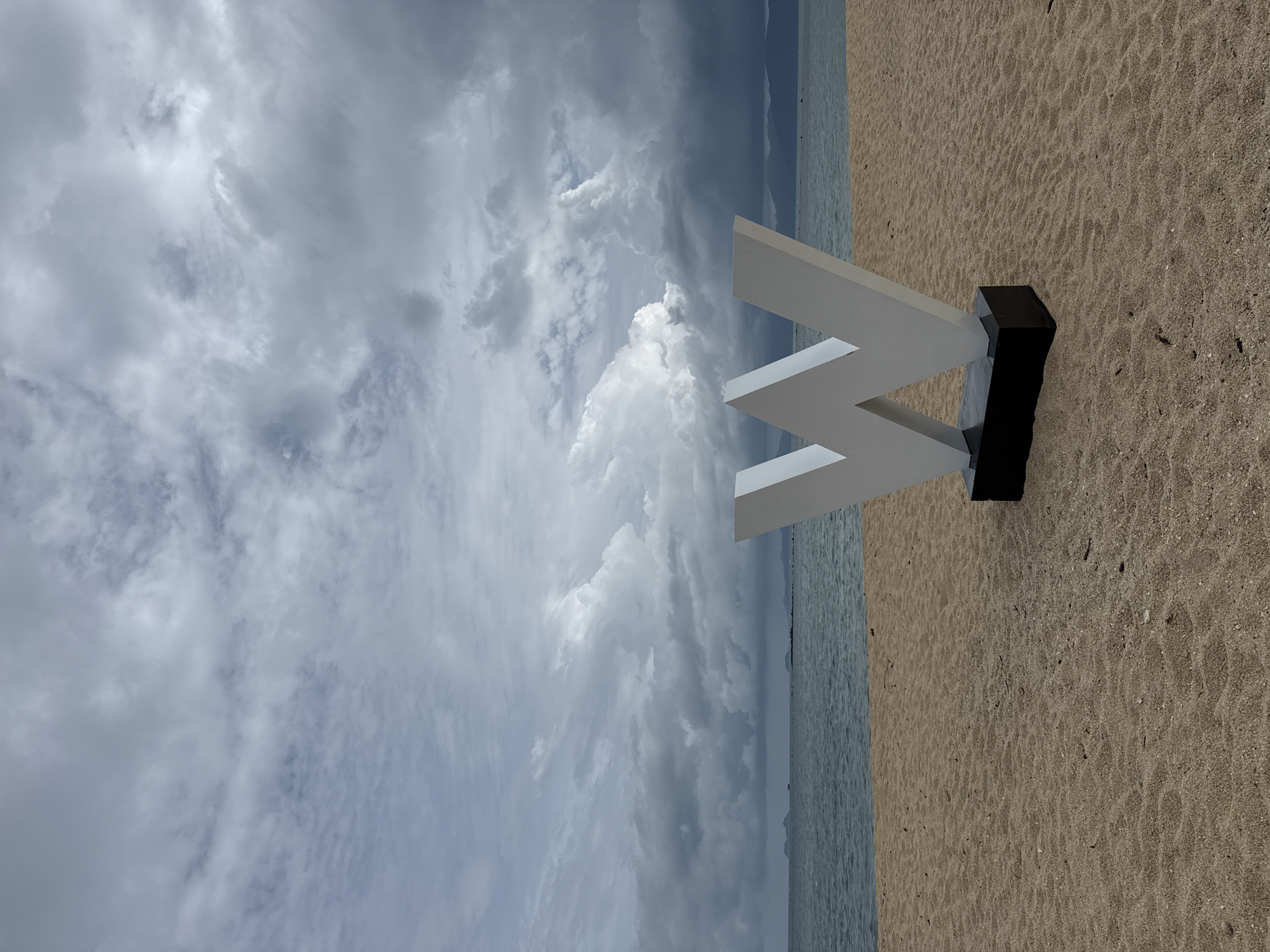 The image depicts a beach scene in the Ko Samui District of Thailand under a cloudy sky. In the foreground, a large, white, three-dimensional letter "W" stands prominently on a black base on the sandy beach. The letter is a significant focal point. Behind the "W," the ocean stretches out, with a distant shoreline visible on the horizon. The sky is filled with dramatic, grey clouds, suggesting an overcast or potentially stormy weather condition. The sand is light brown and appears textured, with some small debris scattered across it. The water is a muted blue-green. The scene is devoid of any people or animals. The overall atmosphere is somewhat somber due to the heavy cloud cover.