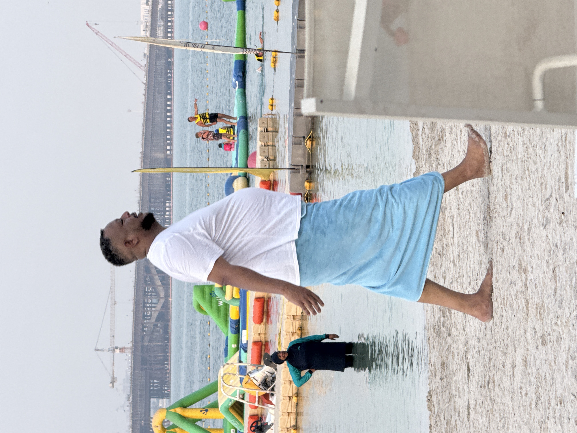 In an outdoor, daytime scene at a beach, a man with a beard, wearing a white t-shirt and a light blue towel wrapped around his waist, walks across the sand. The man is facing right in profile, with his mouth open as if speaking. In the background, a body of water contains inflatable water structures and several people. One person, wearing a dark hijab and swimwear, stands in the shallow water. Further in the background, a large construction project is underway, with cranes and a bridge visible. The sky is overcast.