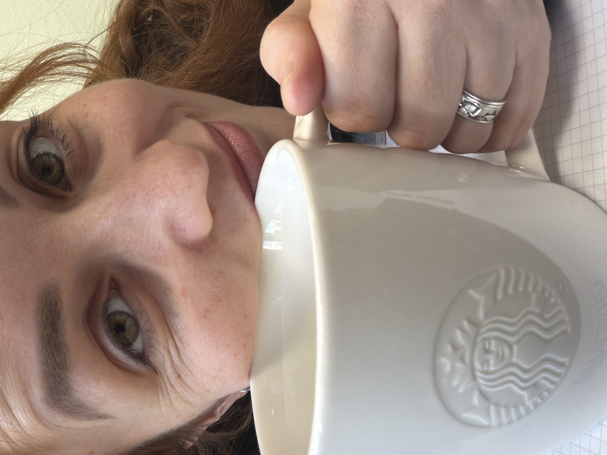 A fair-skinned woman with reddish-brown hair is positioned close to the camera, her face tilted towards the left. She is holding a large white mug, featuring an embossed Starbucks logo, to her lips as if to take a drink. Her left hand grips the mug's handle, and a silver ring adorns her ring finger. The background includes a glimpse of grid-lined paper. There is no indication of the location being the city of Chaikovsky, Russia.