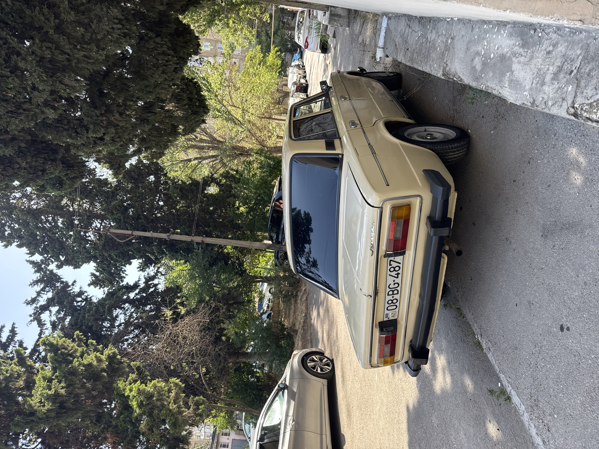 A vintage beige sedan with the license plate "08-BG-487 AZ" is parked on a paved street lined with trees. The car is angled towards the viewer's right, with its rear end prominently featured. The license plate indicates its registration in Azerbaijan. In the background, several other vehicles are visible, along with buildings and lush green trees, suggesting a residential area in Baku. The sun casts shadows on the ground, indicating it is daytime.