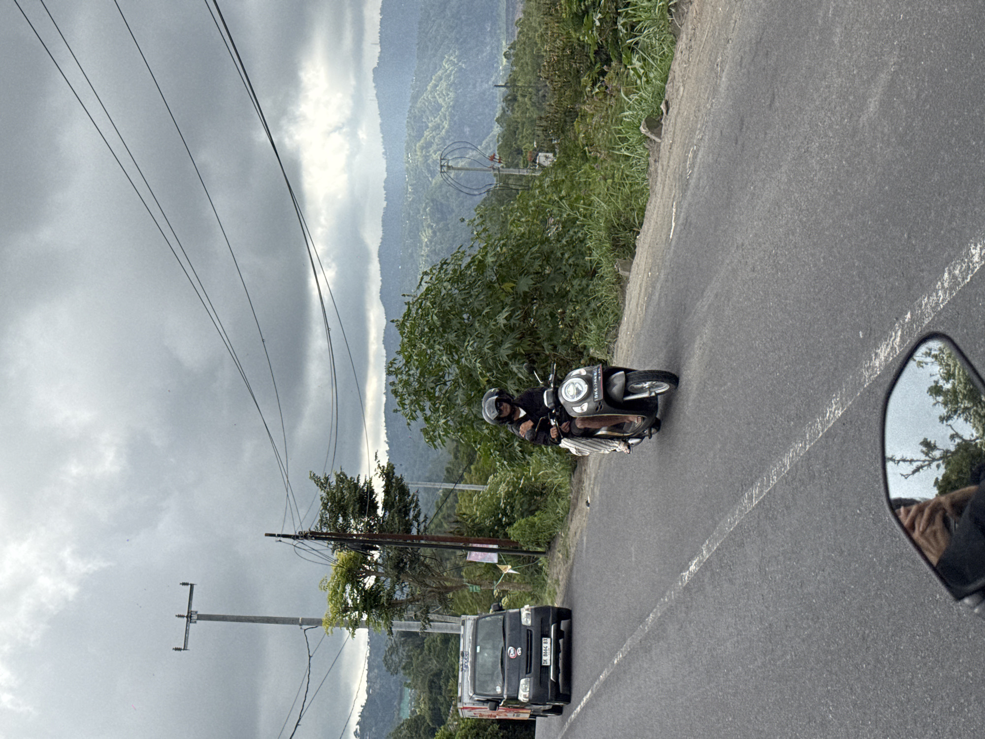 The image depicts a road in Kintamani, Indonesia, with a person riding a scooter and a truck in the background. The sky is overcast with dramatic clouds, and rolling hills covered in greenery form the backdrop. The road is paved and has a white dashed line marking the lanes. A utility pole with wires runs along the side of the road.
On the scooter, a person wearing a helmet and dark clothing is riding. They appear to be a woman, judging by her attire and visible legs. She is wearing a long skirt or dress and sandals. The scooter is dark grey.
In the background, a black truck with a white cargo box is driving in the opposite direction. The truck has license plates that appear to be from Indonesia.
The environment is lush and mountainous, characteristic of Kintamani's landscape. The vegetation is dense, with trees and bushes lining the roadside and covering the hills. The overall atmosphere is slightly muted due to the cloudy weather. The perspective of the shot suggests it was taken from another moving vehicle, possibly a motorbike, as evidenced by the reflection in a mirror at the bottom of the frame.