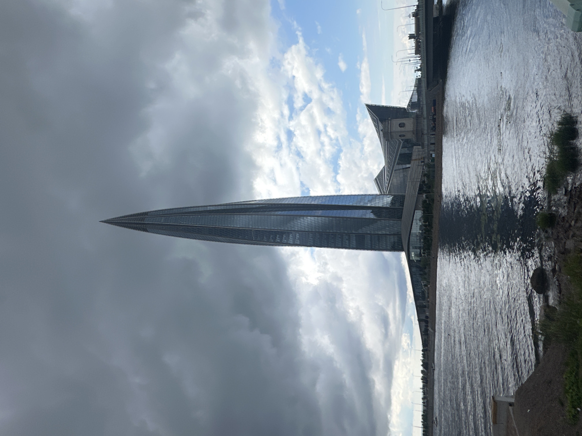 The image captures the Lakhta Center skyscraper, a prominent landmark in Saint Petersburg, Russia, from a waterside perspective. The skyscraper, with its distinctive glass facade and spire, dominates the frame. A body of water, likely part of the Gulf of Finland, occupies the foreground, reflecting the sky and the building. The sky is partly cloudy, with dramatic dark clouds juxtaposed against patches of blue sky.
To the right of the skyscraper, a modern bridge spans the water, with streetlights lining its edge. In the middle ground, a cluster of contemporary buildings with angular roofs forms the base of the Lakhta Center complex. The shoreline in the foreground consists of a mix of gravel and grass, with some rocks visible in the water.
A few small boats are present on the water, and what appear to be people swimming or engaging in water activities are visible in the middle of the water body. The overall scene suggests a contemporary urban landscape at the edge of the city, with a focus on modern architecture and its relationship with the natural environment.