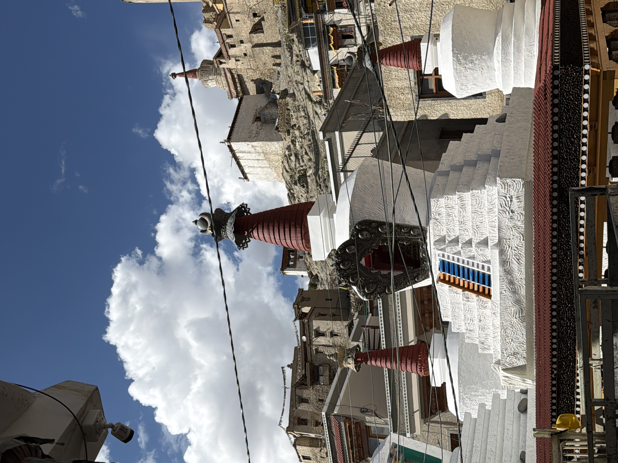 The image depicts a portion of a Tibetan Buddhist monastery situated in the city of Leh, India. In the foreground, there are multiple white stupas, which are dome-shaped Buddhist reliquary monuments. The largest stupa in the center has a tiered base decorated with intricate patterns and a prominent red spire. A blue and orange ladder is placed against its side. To its left, another smaller stupa with a similar red spire is visible. To the right, a third white stupa with a red spire is partially visible.
Behind the stupas, a complex of buildings constructed from stone and whitewashed plaster climbs up a rocky hillside. These structures, with their flat roofs and small windows, are characteristic of traditional Tibetan architecture. Prayer flags, though not clearly discernible, are likely present on the rooftops, as is common in such locations.
In the upper portion of the image, the sky is a vibrant blue with scattered white, cumulus clouds. Several black wires crisscross the frame, indicating the presence of electricity or communication lines. A security camera is mounted on a white structure in the upper left corner of the image. The overall scene conveys a sense of religious and architectural grandeur against a natural mountain backdrop.