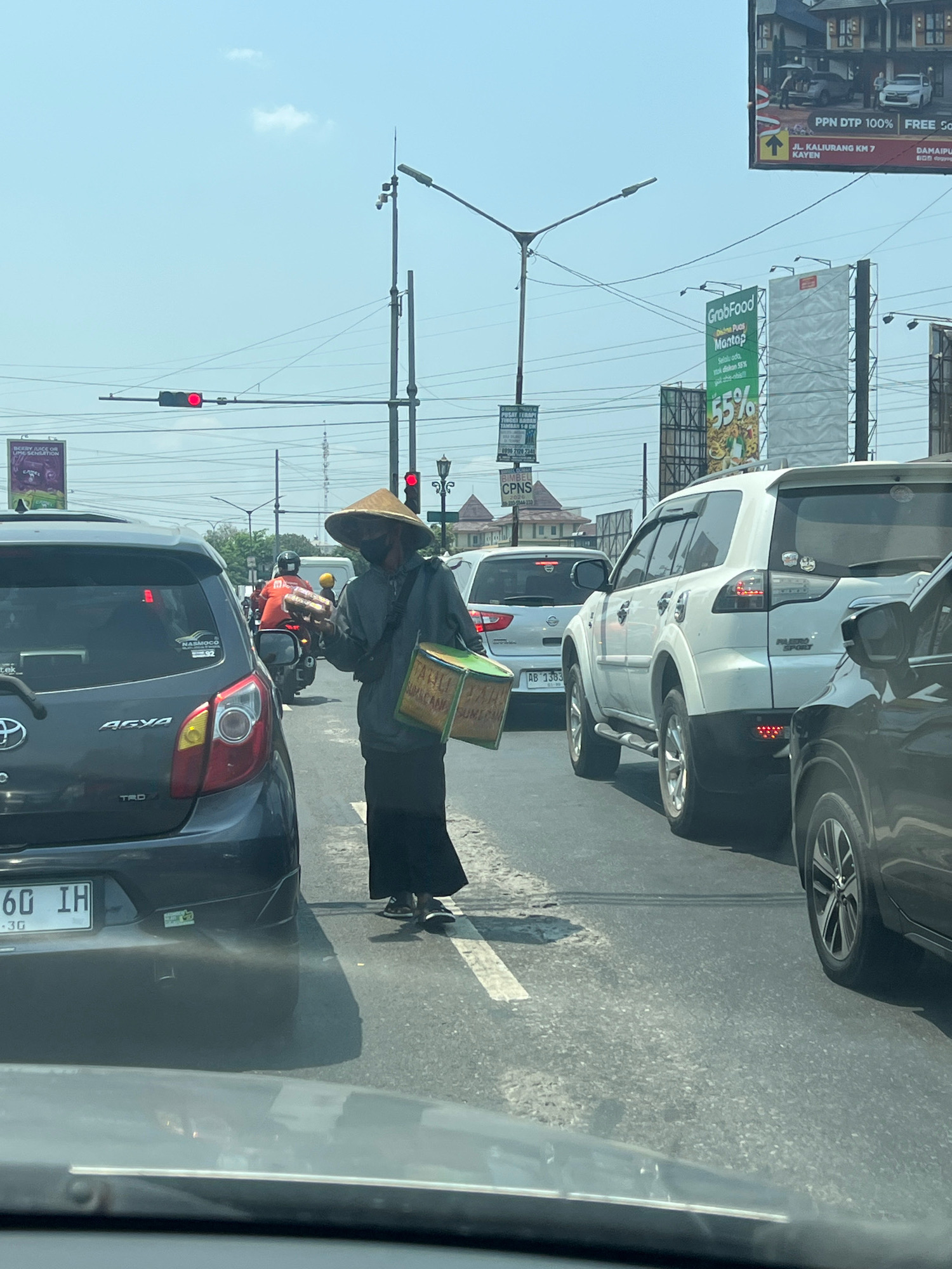 An outdoor, eye-level, medium shot captures a street scene in Ngaglik, Indonesia, on a clear, sunny day. A person wearing a conical hat and a dark long-sleeved shirt and skirt walks across the street, carrying a rectangular green box with yellow lettering on their right shoulder. They are positioned between two lanes of stopped traffic. To the left, the rear of a dark grey car is visible, with a license plate that reads "60 IH". Further back, a white SUV is stopped in the right lane. The background reveals multiple billboards, traffic lights (one red), streetlights, and buildings. The road is asphalt with white lane markings.