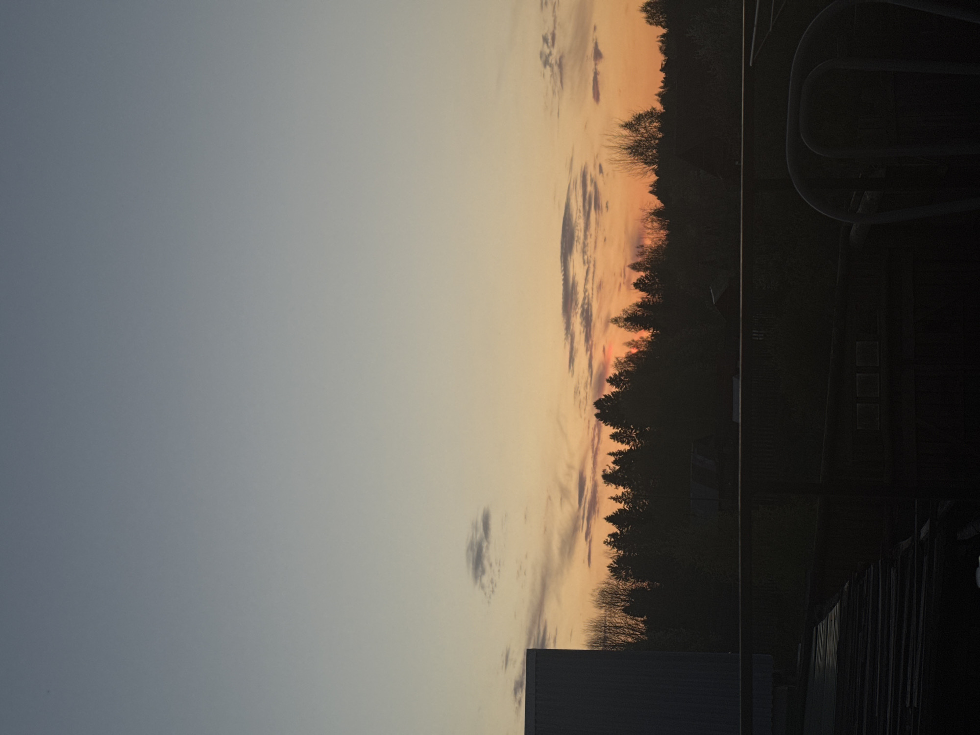 The image shows a sunset sky. The left side of the image shows a light blue sky transitioning into shades of orange and pink near the horizon. Silhouetted trees are visible against the sunset colors. In the bottom right corner, parts of a building and railing are visible, suggesting the photograph was taken from an elevated position within a developed area. The location is Pavlovsky, Russia.