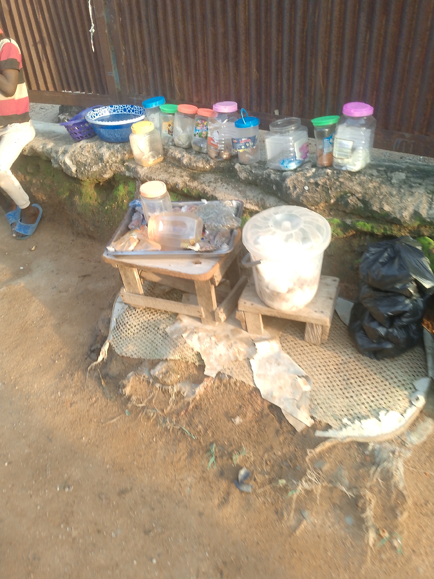 A person wearing a red, white, and black striped shirt and white pants with blue flip-flops stands in the lower left of the frame. They are near a concrete ledge on which several plastic containers are placed. To the right of the person, a blue basket and a blue and white patterned bowl sit on the ledge. Further to the right are multiple clear plastic jars with colored lids, containing various items, and some smaller plastic containers. In the foreground, a small wooden stool holds a metal tray with more containers and a large clear plastic bucket filled with white contents. Next to the stool, a pile of black trash bags is visible. The scene appears to be outdoors, with a corrugated metal wall in the background and a sandy ground in the foreground.