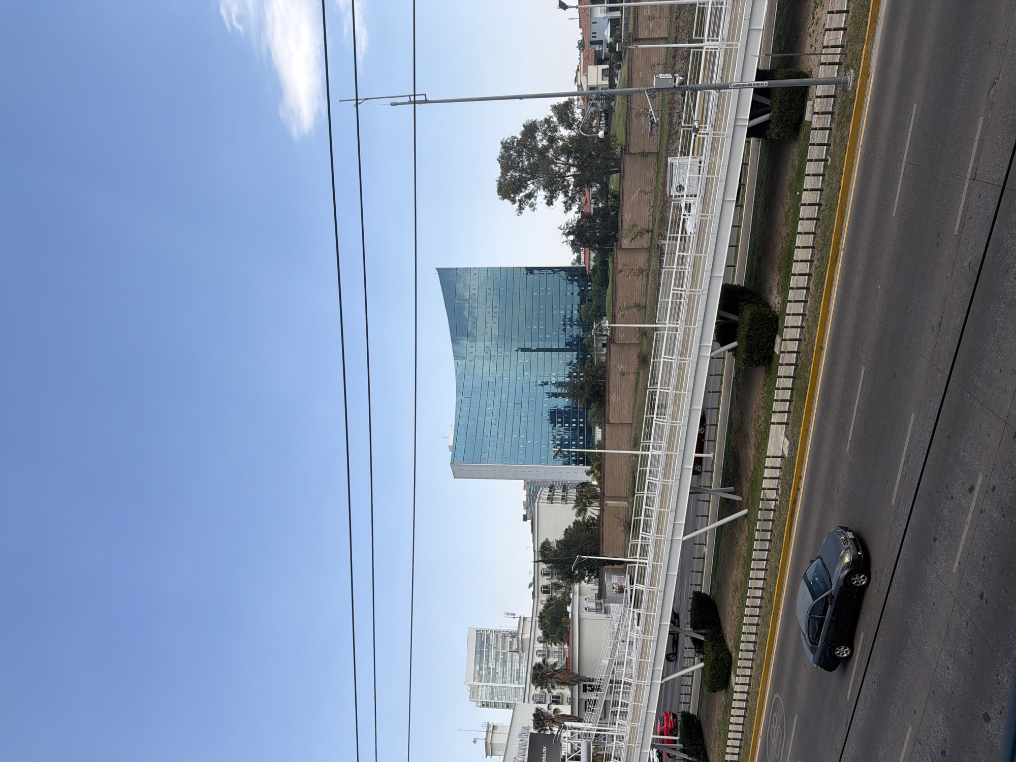 This is an outdoor, elevated view of an urban landscape in San Bernardino Tlaxcalancingo, Mexico, captured on a clear, sunny day. The sky, occupying the upper portion of the frame, is a vibrant blue with a single wispy white cloud visible in the upper right corner. Several dark power lines stretch horizontally across the upper and middle sections of the image.
In the mid-ground, a prominent modern building with a curved, reflective glass facade dominates the left side. Its light blue or teal exterior mirrors the sky and surrounding environment. Behind and around this structure are other buildings and lush green trees, including a large, full tree positioned directly behind the glass building.
A white pedestrian bridge with intricate railings and supports traverses diagonally across the scene, appearing to span over a multi-lane asphalt road. The road, located in the lower part of the image, features distinct yellow curb lines and white dashed lane markers. Concrete blocks or planters run alongside the road, separating it from the area beneath the pedestrian bridge.
One dark-colored sedan is clearly visible on the road in the lower right, traveling towards the right side of the frame. Other vehicles, including a partially visible red car, are present further down the road or in the background. Streetlights and poles are interspersed throughout the scene; one pole near the pedestrian bridge displays a sign that reads "SISTEMA DE ALERTA INTELIGENTE" (Intelligent Alert System). Below this, smaller text includes "KANOKEE" and "www.uam.edu.mx". The image depicts a typical, active urban environment with modern infrastructure under bright daylight.