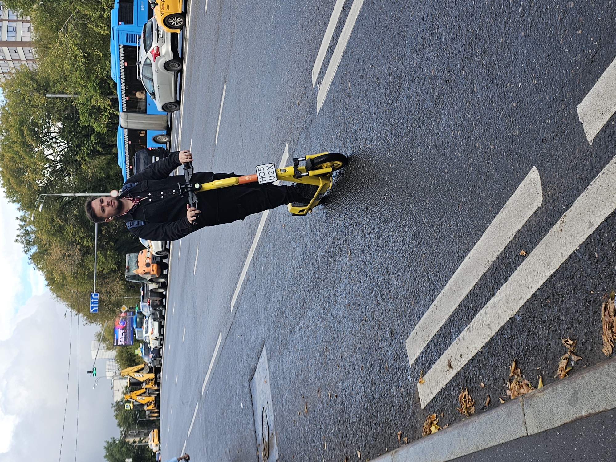 A young man with a beard rides a yellow electric scooter down a wet city street in Moscow, Russia. He wears a black jacket, dark pants, and black shoes. A blue backpack is visible on his back. The scooter has a license plate with the characters "HO5 02X". The street is wet, indicating recent rain, and has white lane markings. In the background, several vehicles are present, including a blue bus, cars, and construction equipment, such as yellow backhoe loaders. Trees line the side of the road, and a tall building is visible in the distance. The sky is overcast.