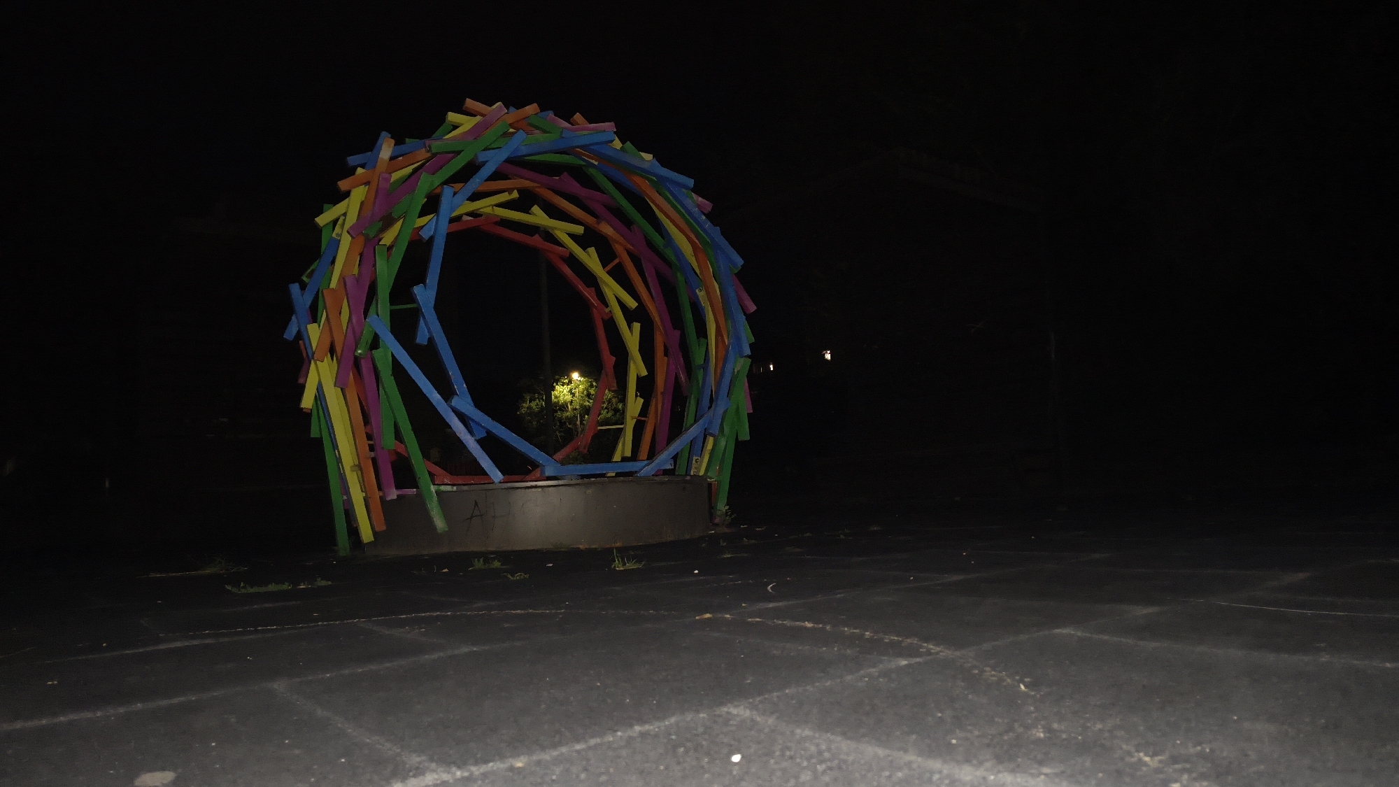 A brightly colored, circular sculpture made of wooden sticks is the focal point of this nighttime image. The sculpture is composed of numerous sticks in shades of red, yellow, green, blue, and pink, arranged in a circular, tunnel-like formation. It sits on a dark, circular base, possibly a planter or pedestal, with a metallic finish. The sculpture is lit from within by a warm yellow light, revealing glimpses of greenery and foliage behind it. The ground surrounding the sculpture is dark and textured, appearing to be a paved surface, possibly rubberized, with some scattered debris. The background is entirely black, indicating darkness and a lack of ambient light, with the exception of a faint, distant light source on the right side of the frame. The overall scene suggests an outdoor public space, such as a park or playground, in Mikhaylovka, Russia. There are no people or animals visible in the image.