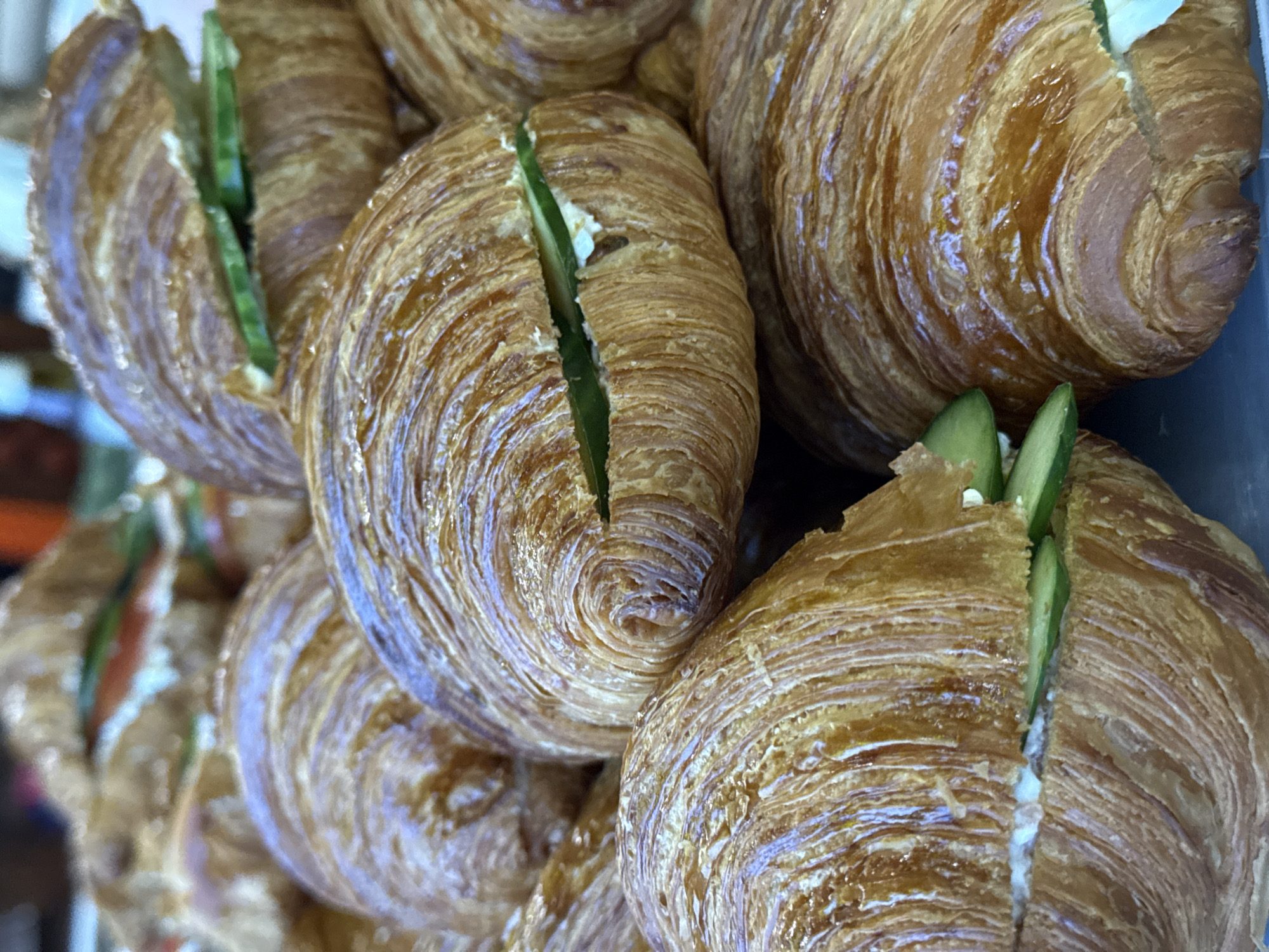 The image shows a close-up of a pile of croissants. Each croissant has a vertical slice through the middle, and a few slices of cucumber are inserted into the cut. A white spread is also visible inside the croissants, likely cream cheese or a similar filling. The croissants are golden brown and appear freshly baked. The image was taken in Perm, Russia.