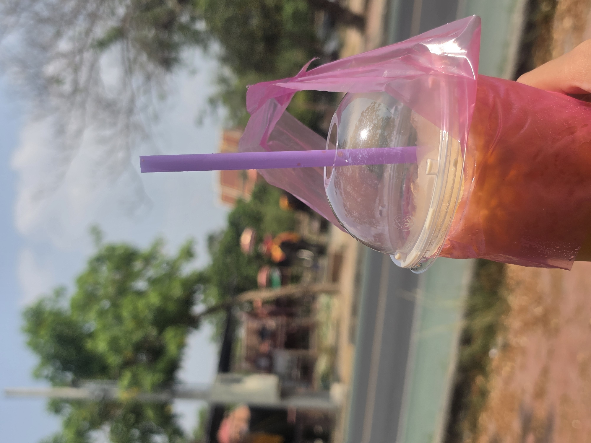 The image captures a close-up of a vibrant beverage being held outdoors, likely in Phra Nakhon Si Ayutthaya, Thailand, given the context. The main subject is a clear plastic cup filled with a reddish-orange or pinkish-red drink, possibly a fruit smoothie or iced tea. A clear domed lid covers the cup, from which a bright purple plastic straw protrudes. The entire cup is encased in a translucent pink plastic bag, a common way to carry drinks in some Asian countries. A hand is visible holding the drink from the right side of the frame.
The background is softly blurred, creating a shallow depth of field that keeps the focus sharply on the beverage. It depicts an outdoor scene under bright daylight, suggesting a warm and sunny day. Visible elements in the background include lush green trees, a clear blue sky with some wispy white clouds, and a faint outline of a paved road or pathway. Distant, blurred structures with reddish-orange roofs can be discerned through the foliage, consistent with local architecture. No discernible text is visible on the cup, bag, or straw. The overall impression is a refreshing drink enjoyed casually outdoors.