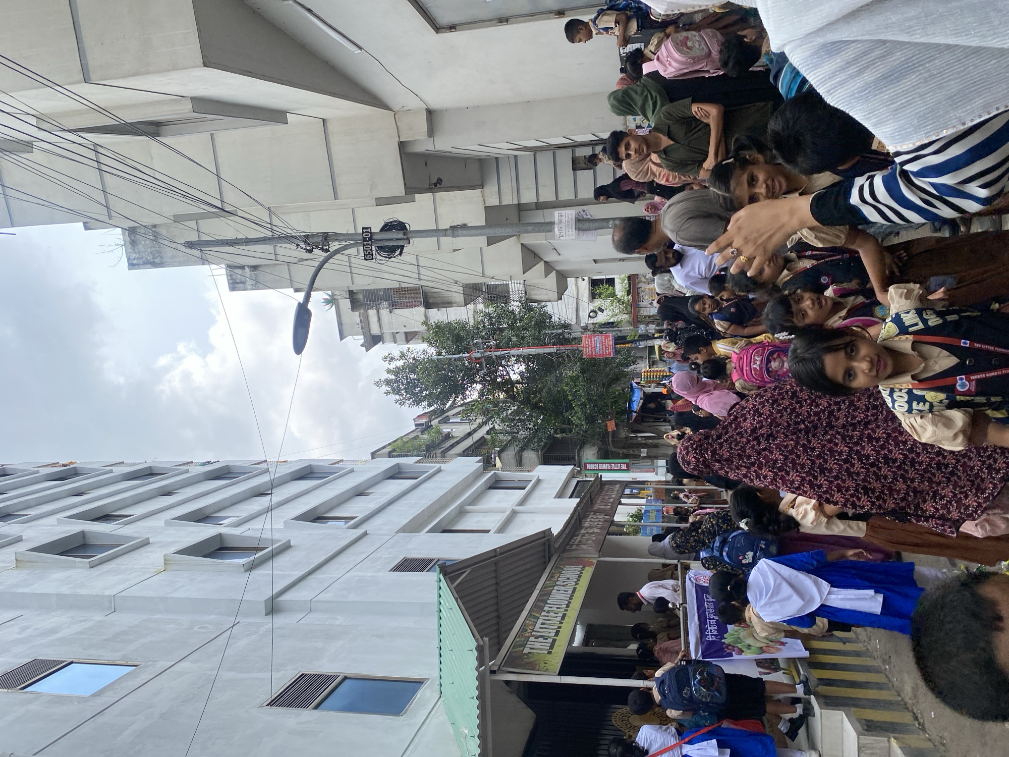 A group of people, mostly children wearing school uniforms, are gathered outside "The Little Flower School" in an urban setting. The school appears to be a multi-story building. Many children have backpacks. The sky is overcast. Some adults are also present, including parents and school staff. There are power lines and streetlights visible, indicating a city environment. A banner for the school is displayed prominently. Some individuals are wearing hijabs and burqas. The scene suggests a school day commencement or dismissal.