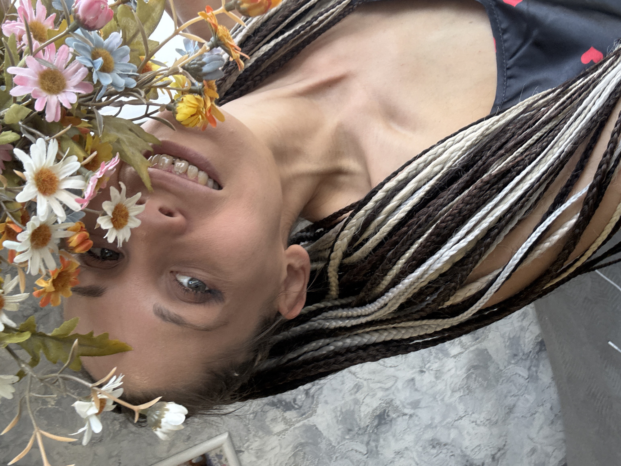 The image is a portrait of a woman with braided hair, featuring alternating dark brown and blonde strands. She is wearing a black top with a pattern of red hearts. The woman is smiling, and a bouquet of colorful artificial flowers is positioned near her face. The background appears to be a gray textured wall. The location is in the city of Fedorino, Russia.