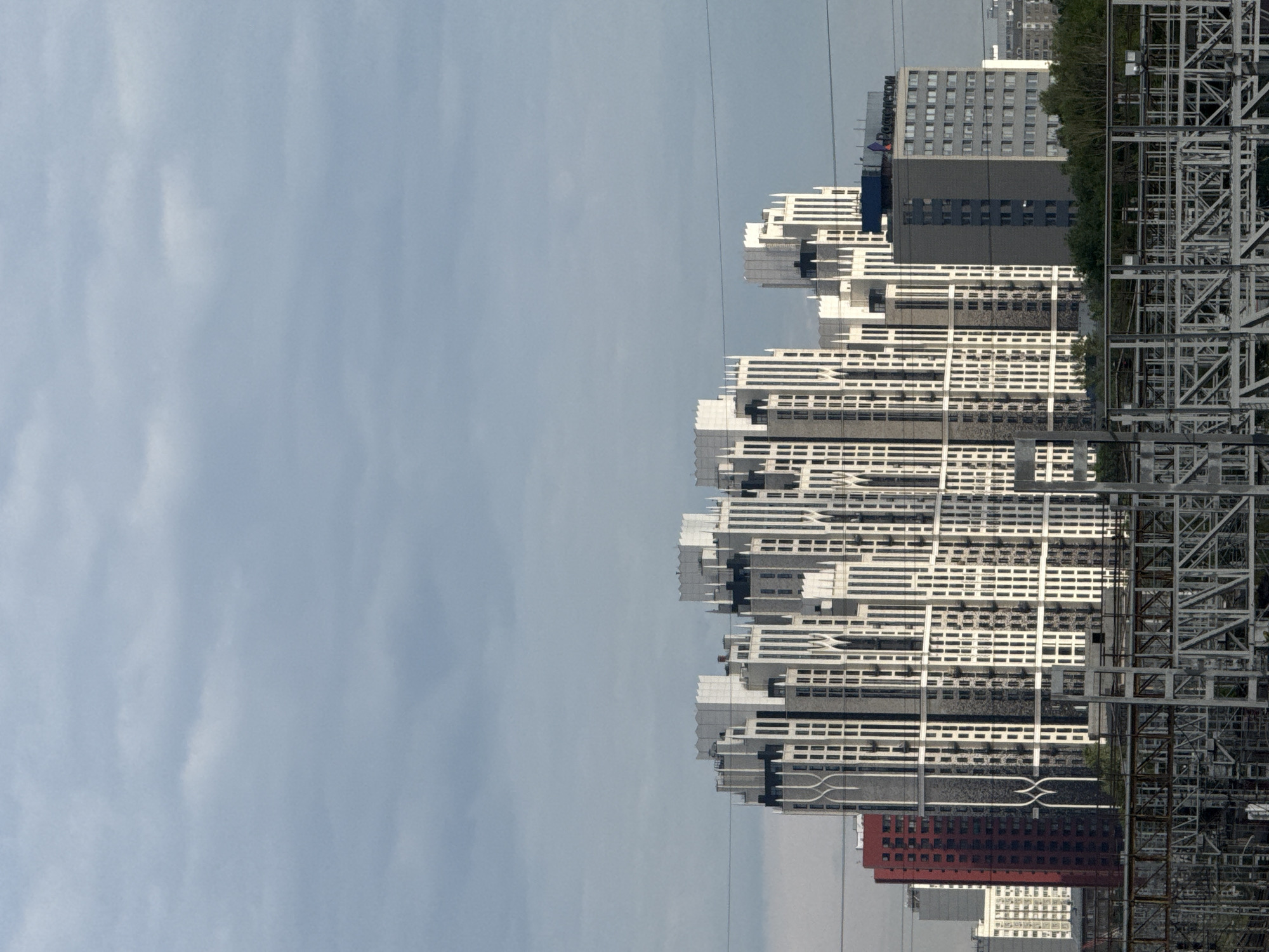 The image shows a skyline of modern residential buildings in Moscow, Russia, under a cloudy sky. In the foreground, there is an intricate metal structure, possibly part of a railway or industrial facility, with power lines running across the frame. The dominant feature of the mid-ground is a complex of tall, white and grey apartment buildings with an ornate, stepped architectural style. To the left of this complex, a bright red building stands out. To the right, a grey office building with the logo "Ростелеком" (Rostelecom) is visible. Lush green trees line the bottom of the image behind the metal structure. The scene appears to be captured during daylight, with diffused light from the overcast sky.