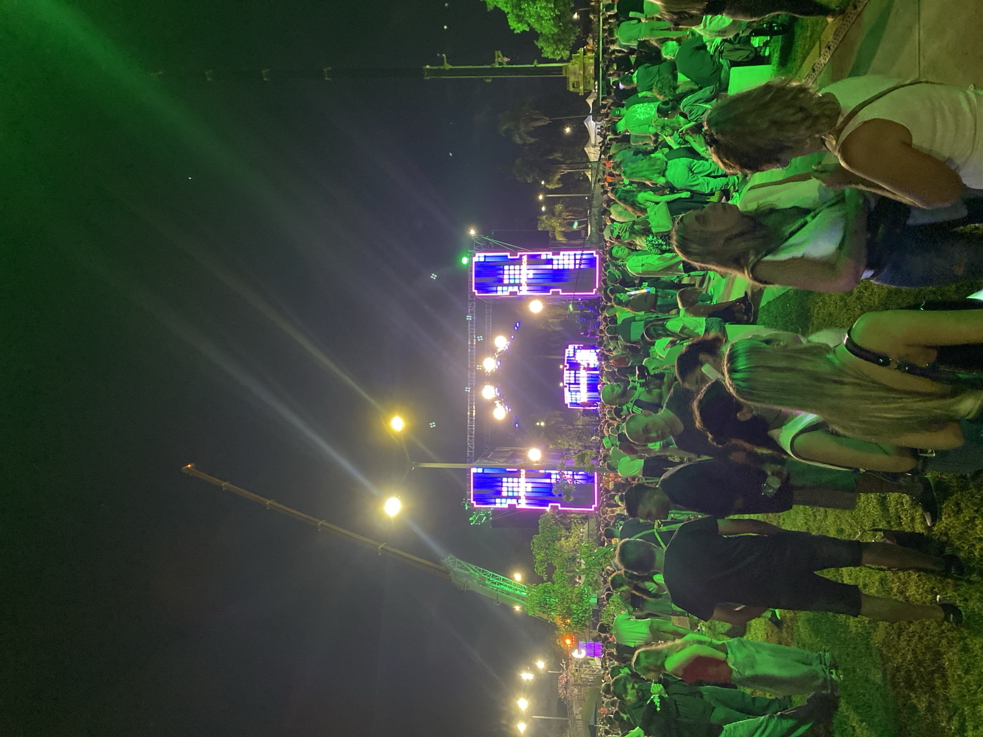 Here's a description of the image:
The image captures a nighttime outdoor event in what appears to be a public space. A large crowd of people is gathered, illuminated by green lighting. In the background, a stage with two large, brightly lit screens featuring pixelated designs dominates the scene. Streetlights are interspersed throughout the crowd and near the stage. A crane extends into the sky on the left side of the frame.
Although the city and location are not explicitly confirmed in the image, the scene resembles a large public gathering, potentially a concert or festival, at night. Without specific landmarks or identifying features, confirming it is Buenos Aires, Argentina, is impossible based on the image alone.