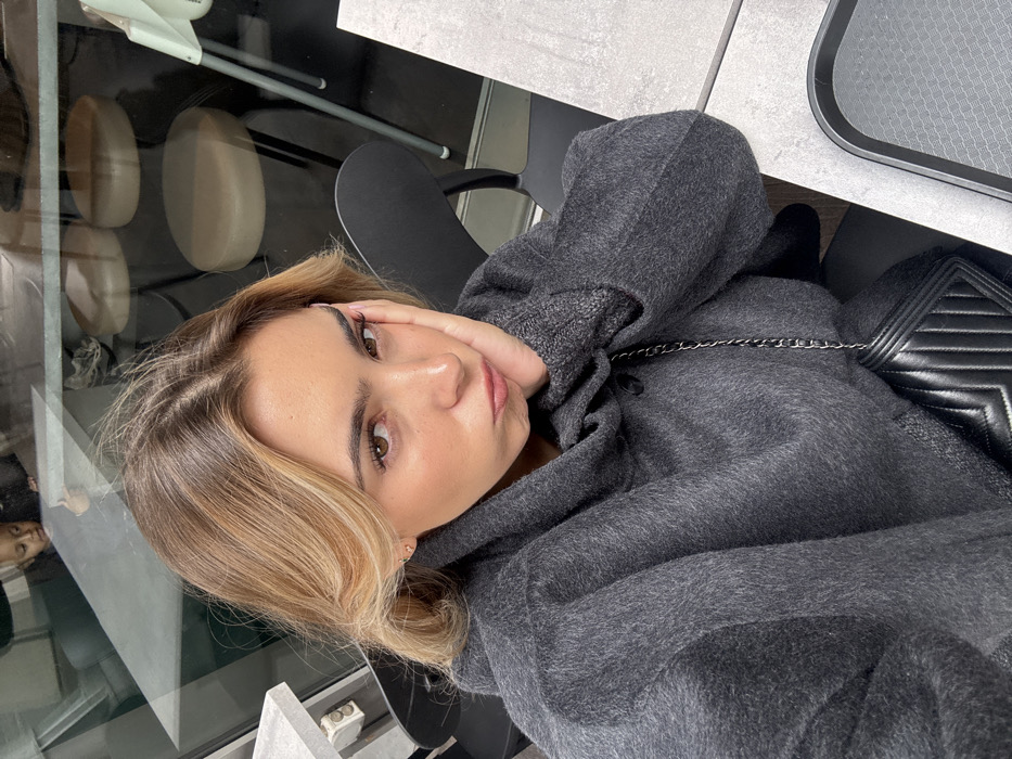 A young adult female is depicted in a medium-close-up selfie, seated at a light-gray table within an indoor commercial establishment, likely a cafe or restaurant, located in Vaulx-en-Velin, France. The subject has blonde hair with darker roots, brown eyes, and is wearing a dark charcoal-gray, textured, hooded garment. Her right hand (viewer's left) rests against her cheek, and her lips are pursed in a pout, directed towards the camera. A black quilted handbag with a silver-colored chain strap is visible across her body. On the table to her right (viewer's left), a black serving tray is present. The background consists of reflections in a glass panel, revealing elements of an interior space, including a young child with light hair looking away from the camera, two round light-colored stools, and a light-colored high chair.