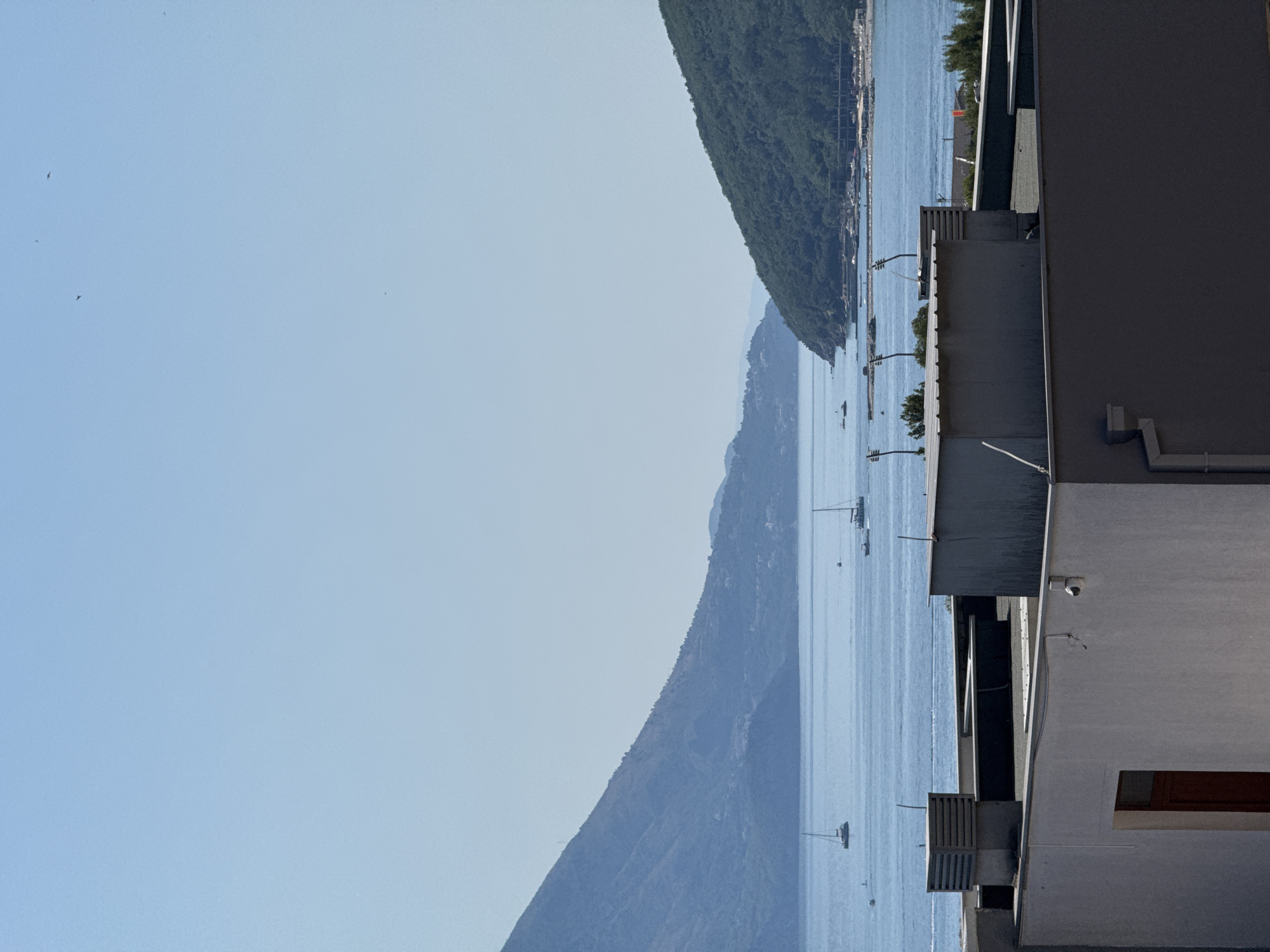 This is a vertical, eye-level shot of a body of water with a clear blue sky above. In the foreground, a modern building's flat rooftop occupies the lower third of the frame, featuring several vents and architectural details. Beyond the rooftop, the calm blue water stretches to the horizon, dotted with several sailboats and a few smaller boats. On either side of the water, lush green mountains rise, partially shrouded in a light haze, suggesting a warm, possibly humid, climate. The scene is tranquil and serene, evoking a sense of peace and natural beauty. This vista is consistent with views from Budva, Montenegro, a coastal town known for its stunning Adriatic coastline and mountainous backdrop.