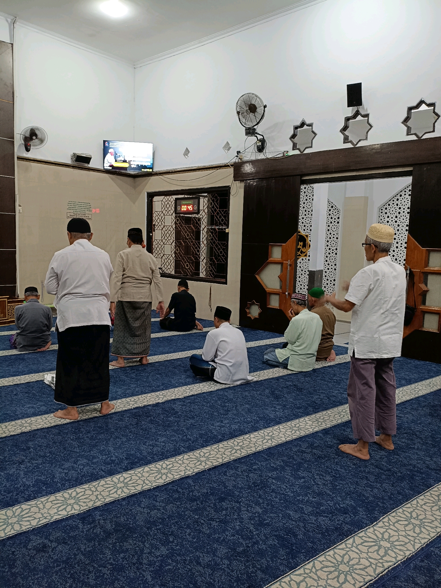 In Maguwoharjo, Indonesia, within a mosque, several men are engaged in prayer. The scene is captured from a mid-level perspective, looking towards the rear of the prayer hall. A large, dark brown wooden structure with ornate geometric patterns dominates the background, serving as a doorway or partition. Above and to the left, a television screen displays a religious broadcast, and a digital clock shows "00:45".
The floor is covered in a deep blue carpet with a repeating light-colored floral pattern forming parallel strips. On the carpet, men are positioned in rows, facing the front of the mosque, in various stages of prayer. Some are seated with their knees bent and legs tucked beneath them, while others are standing or in a kneeling position.
A man in the foreground, on the right side of the frame, is standing and gesturing with his right hand. He is wearing a light-colored long-sleeved shirt, a light-colored cap, and purple trousers. His feet are bare, as are the feet of most other men in the prayer hall.
To the left of the standing man, another man is seated, facing away from the camera, wearing a white long-sleeved shirt and a black sarong. Further to the left, another man in a grey shirt and black sarong is also seated.
The walls are painted a light color. A fan is mounted on the wall to the left, and several decorative, star-shaped cutouts are visible along the top of the dark wooden structure. The lighting is bright, emanating from overhead sources, creating a well-lit interior.