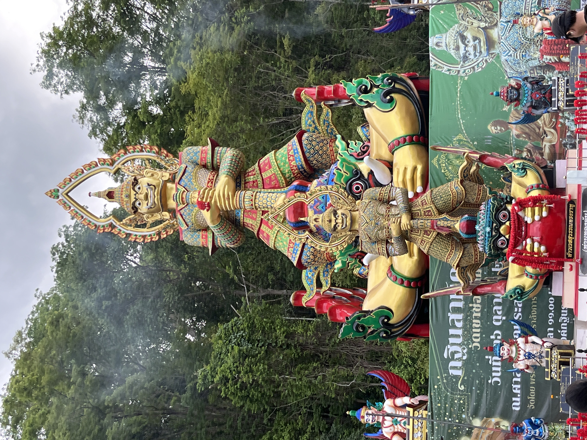 The image depicts a religious display, likely related to the Kathina ceremony, possibly located in or near Chao Phraya Surasak, Thailand, given the Thai script on a banner in the background. Two figures are prominent: a large golden statue of a humanoid figure, possibly representing a deity or mythological being, with intricate decorative elements. A smaller, equally ornate figure rests beneath the larger one. These figures are likely part of a larger procession or display, indicated by additional figures and decorative elements visible at the edges of the frame. The background includes lush green foliage and trees, suggesting an outdoor setting, possibly a temple ground or public space. Smoke is visible in the upper portion of the image, possibly from incense or other ceremonial activities. The overall scene appears vibrant and colorful, indicative of Thai religious and cultural celebrations.