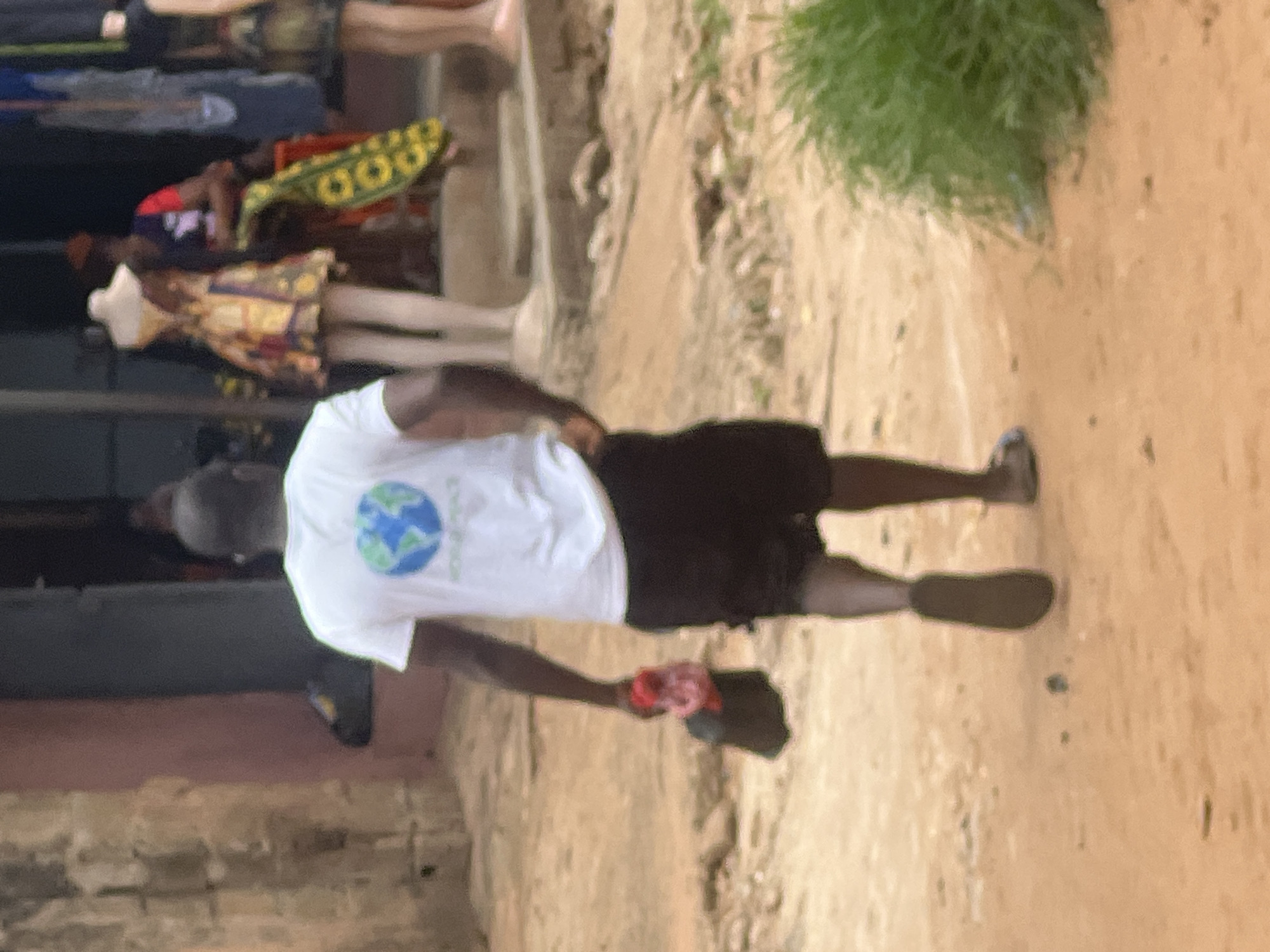 An outdoor shot shows a man in a white t-shirt and black shorts walking on a dirt path. The man holds a hat or cloth in his right hand. In the background, people stand near a structure. Green foliage grows near the path. The location is Unknown, Unknown.