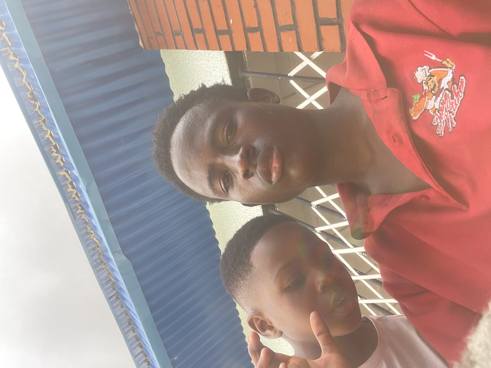 The image captures a close-up, selfie-style shot of two young Black males outdoors. The individual in the foreground, wearing a red polo shirt with an embroidered logo of a cartoon chef holding a fork and a plate of food, is positioned to the right. His face is turned towards the camera, and he has short, dark, curly hair. To his left and slightly behind him, another young Black male is visible, with short hair and wearing a light-colored t-shirt. He is looking towards the camera and holding up his left hand, with his index finger and thumb forming a circle.
The background reveals elements of an urban environment. A blue, corrugated metal roof is prominent above the subjects, with a section of orange brick wall to the right. Behind the two individuals, a white metal grate or security bars can be seen. The lighting suggests an overcast day.
Given the presence of distinctive red brick architecture, corrugated metal roofing, and what appear to be typical urban street elements in Nigeria, the location is likely within the city of Jos, Nigeria.