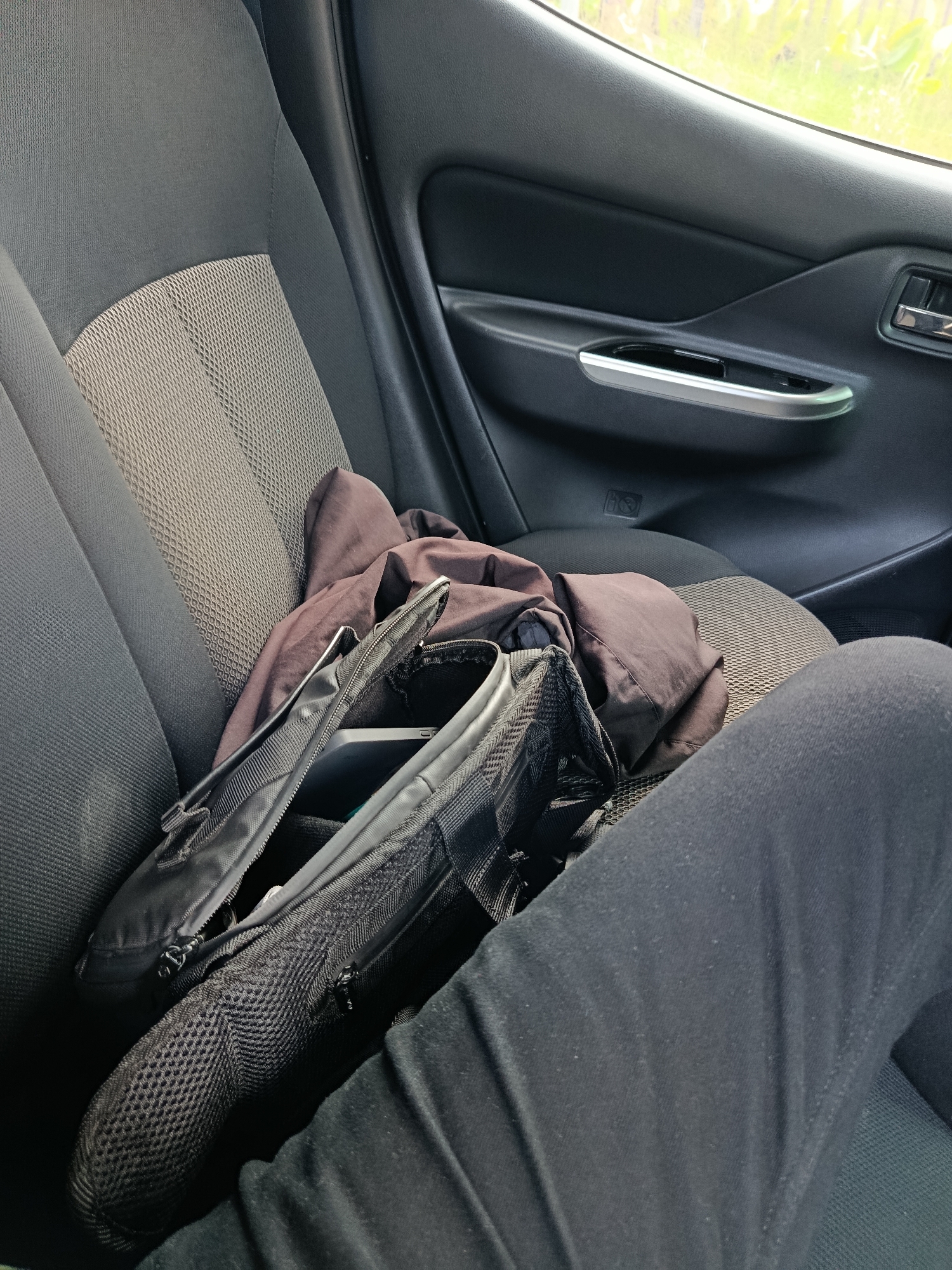 A close-up shot captures a person's legs in black pants, resting on a car seat. A black backpack is placed on the seat, partially open, revealing a glimpse of a phone and a brown jacket inside. The car interior, with its dark upholstery and a silver door handle, suggests a private vehicle. The background, though blurred, shows greenery, hinting at an outdoor or semi-urban setting. The scene is devoid of any specific landmarks that would indicate its location in Cisarua, Indonesia.