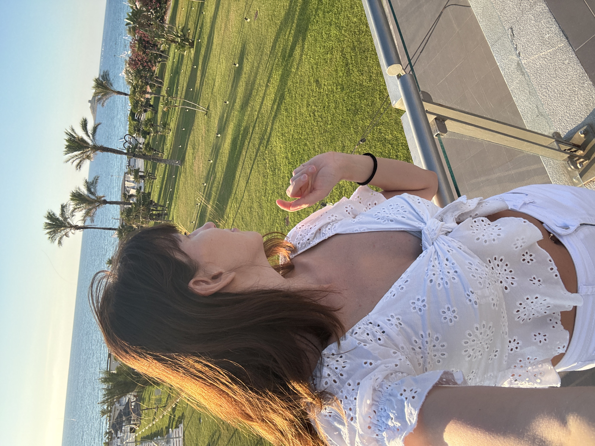 A woman stands on a balcony overlooking a resort area with a view of the sea in Isola delle Femmine, Italy. The woman has brown hair and is wearing a white eyelet crop top tied at the front and white pants. She is looking to her right, with her left arm bent at the elbow and her hand near her face. A black band is visible on her wrist. The balcony railing is made of metal and glass. In the background, there is a well-maintained green lawn with scattered string lights, palm trees, and other vegetation. Beyond the lawn, the blue sea stretches to the horizon under a clear sky. The sun is casting long shadows across the lawn, indicating it is late afternoon or early evening.