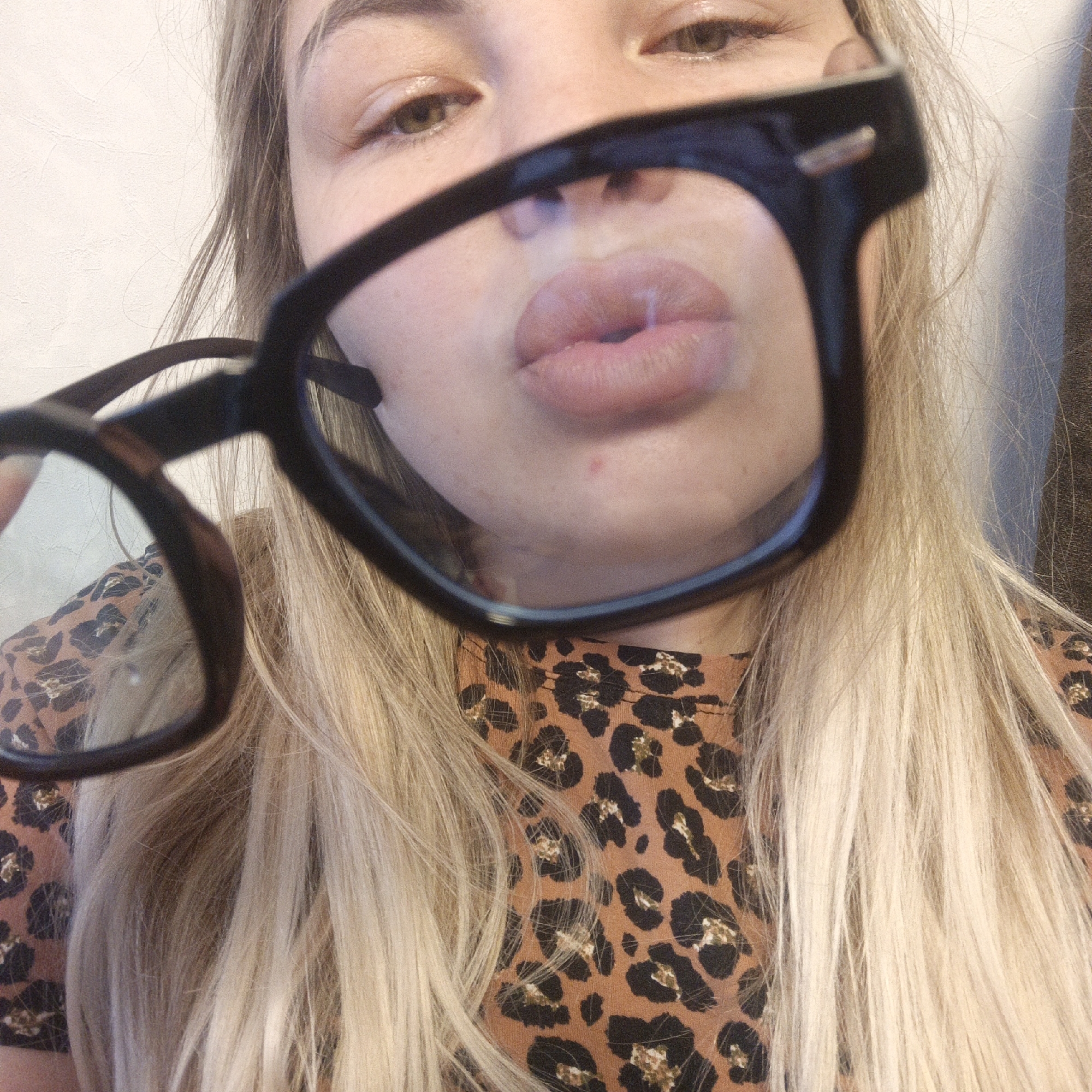 A close-up, high-angle indoor shot features a person, likely female, with long blonde hair, holding a pair of black-framed eyeglasses up to their face. The perspective is from above, looking down towards the person's mouth and nose through the lenses, while their eyes are visible just above the frames.
The person's lips are pursed as if blowing a kiss or making a duck face, prominently framed and slightly distorted by the eyeglasses' lenses. Their light brown or hazel eyes are visible above the top rim of the glasses. Long, straight, light blonde hair cascades down around their shoulders, partially obscuring a brown and black leopard print top. The background is a plain, light-colored wall with some subtle texture. The overall impression is a playful and intimate selfie. No text is visible in the image.