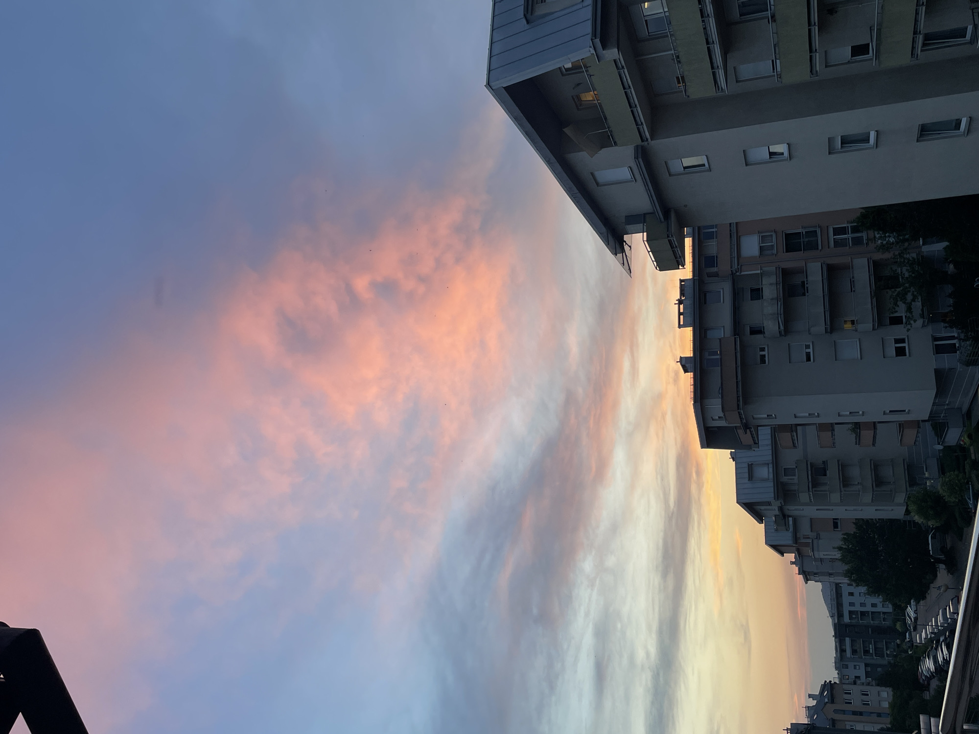 The image depicts a city skyline at dusk. The sky is filled with colorful clouds, displaying hues of pink, orange, and blue, suggesting a sunset. The buildings are tall and modern, with balconies and numerous windows. Some windows appear to be lit, indicating people are inside their apartments. The architecture suggests an urban residential area. The perspective is from a higher vantage point, looking down or across the buildings. There are hints of trees and parked cars visible on the street below. The overall mood is serene, with the warm colors of the sunset creating a peaceful atmosphere. The orientation of the image is rotated 90 degrees counterclockwise.