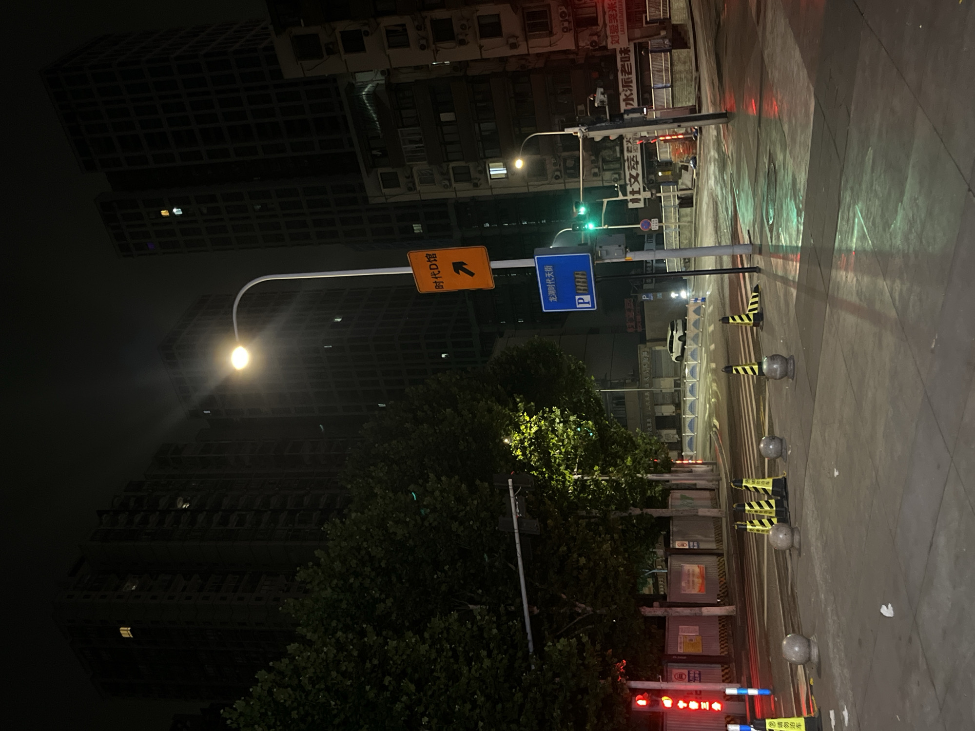 The image depicts a nighttime street scene in the Yuzhong District of China. Tall, dark residential buildings dominate the background, with some windows illuminated. A street lamp with a white curved pole shines brightly. In the midground, there is a traffic light showing red, and various street signs are visible, including an orange sign with Chinese characters and an arrow pointing right, and a blue sign with a white "P" and Chinese characters indicating parking. Yellow and black striped traffic cones and gray spherical bollards are placed along the sidewalk and crosswalk. The wet pavement reflects the colorful lights from the traffic signals and streetlights, creating a slick and illuminated surface. Trees with dense green foliage line the left side of the street. A white car is parked on the right side of the road, and several businesses with illuminated signs are visible on the right side of the street. Construction barriers are present in the background.