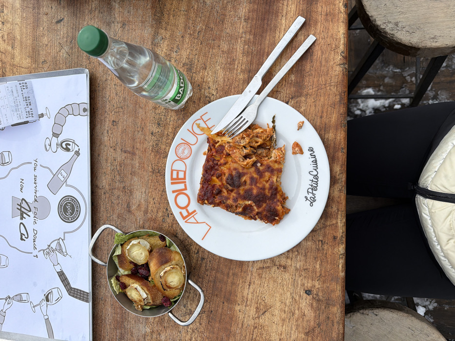 An overhead view captures a meal served on a rustic wooden table at La Folie Douce, Méribel Mottaret, 73550 Les Allues, France. The scene is outdoors, indicated by snow visible on the ground beneath the table and a wooden stool.
On the table, a white ceramic plate, branded "LA FOLIE DOUCE" and "La Petite Cuisine," holds a partially consumed slice of baked pasta, resembling lasagna, with a browned, crispy top. A silver fork and knife rest on the plate's right side. To the upper left of the plate, a small, silver-toned metal pot with handles contains four individual servings of what appear to be toasted bread rolls topped with round slices of goat cheese, garnished with green leaves and red cranberries. A clear plastic bottle of Badoit mineral water with a green cap stands to the right of the plate.
Above the food items, a laminated placemat or menu is visible, featuring black and white illustrations of hands holding wine glasses and bottles, with the text "You survived Folie Douce ?" and "Mon Chéri." A partially obscured white receipt, confirming "LA FOLIE DOUCE" and the Méribel Mottaret, Les Allues location, is tucked under the placemat. The lower body of an individual, wearing black pants and a light-colored quilted jacket, is visible seated at the table, suggesting a first-person perspective. The wooden table surface is worn with visible grain and slight indentations.