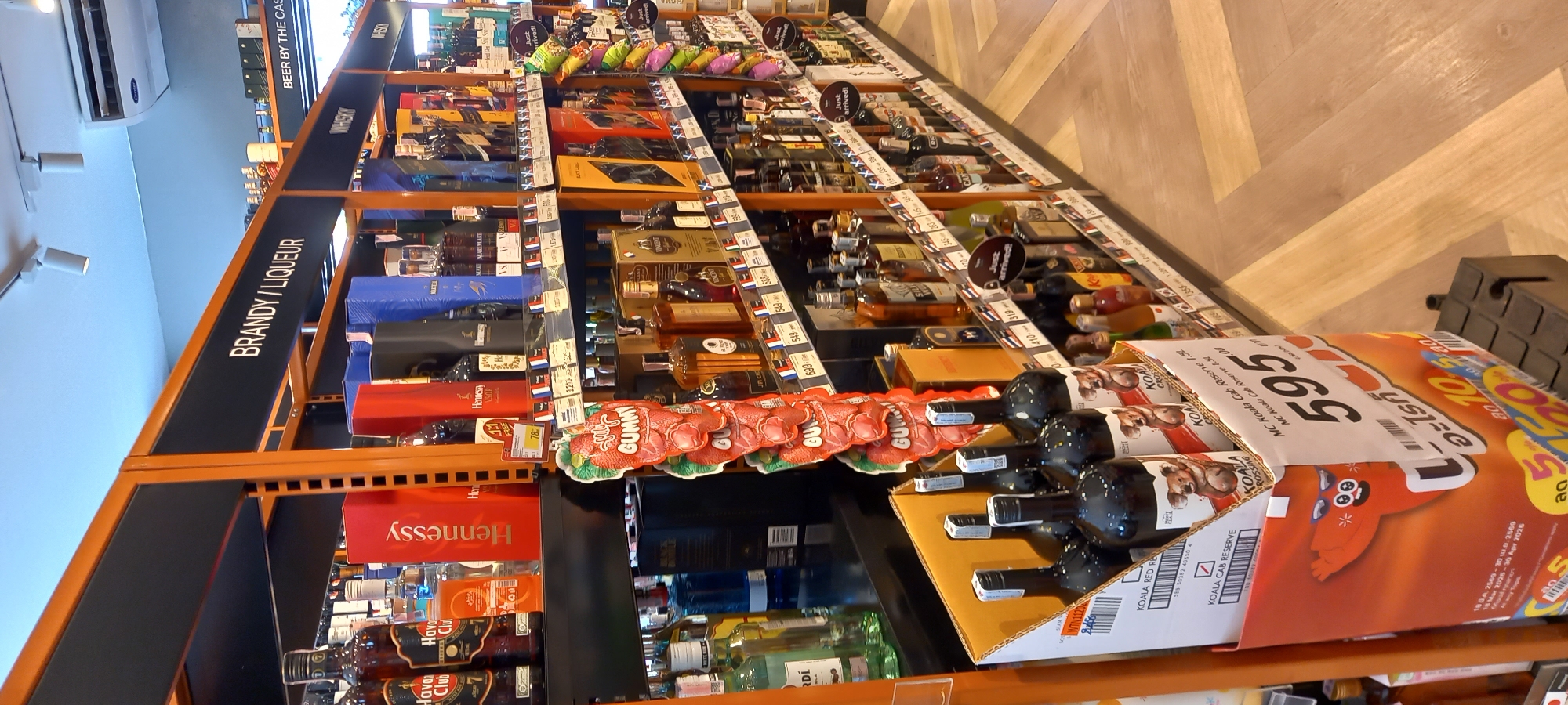 The image displays an aisle in a retail store, likely a liquor store or supermarket, filled with various alcoholic beverages. The shelves are a combination of orange and black, vertically aligned and stocked with numerous bottles. Overhead signs categorize the products, with "BRANDY / LIQUEUR" clearly visible, and "WHISKY" and "BEER BY THE CAS" partially seen further down the aisle.
Specific brands of spirits are identifiable, including "Hennessy VSOP", "Martell" (partially visible), and "Havana Club". A prominent display box contains "KOALA" branded wines, with labels such as "KOALA CAB RESERVE" and "KOALA ROSSINA". Many small price tags are attached to the shelves, showing various numbers like "78", "32", "549", "699", and a larger tag with "595". Promotional text such as "Just arrived!" and "GUMMY" are also present. The KOALA wine box features a cartoon character and Thai text including "อะไรก็" and "ลด" (meaning 'discount' or 'sale'), along with dates "14 Mar 2025 - 30 Apr 2026" which likely refer to a promotional period.
The setting is indoors, with an air conditioning unit and ceiling lights visible. The floor has a light-colored wooden herringbone pattern. The scene depicts a typical retail environment, with products displayed for sale. Given the presence of Thai text and the provided location context, the store is situated in Moo 1, Thailand.
